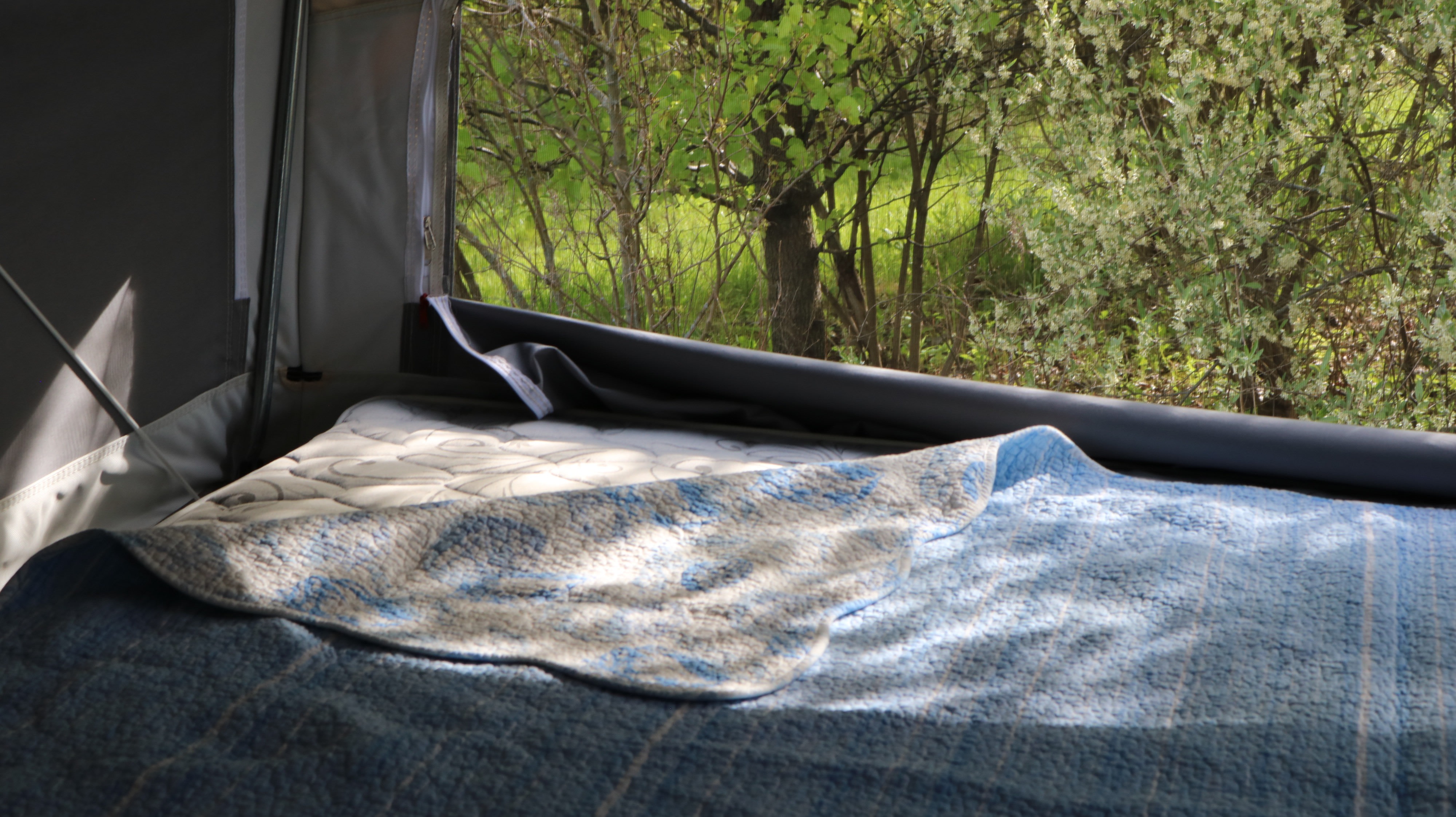 A sunlit bed with blue blankets inside an RV rental, providing a peaceful window view of lush green trees.. Palomino Solaire Hybrid 3 queen beds 2019