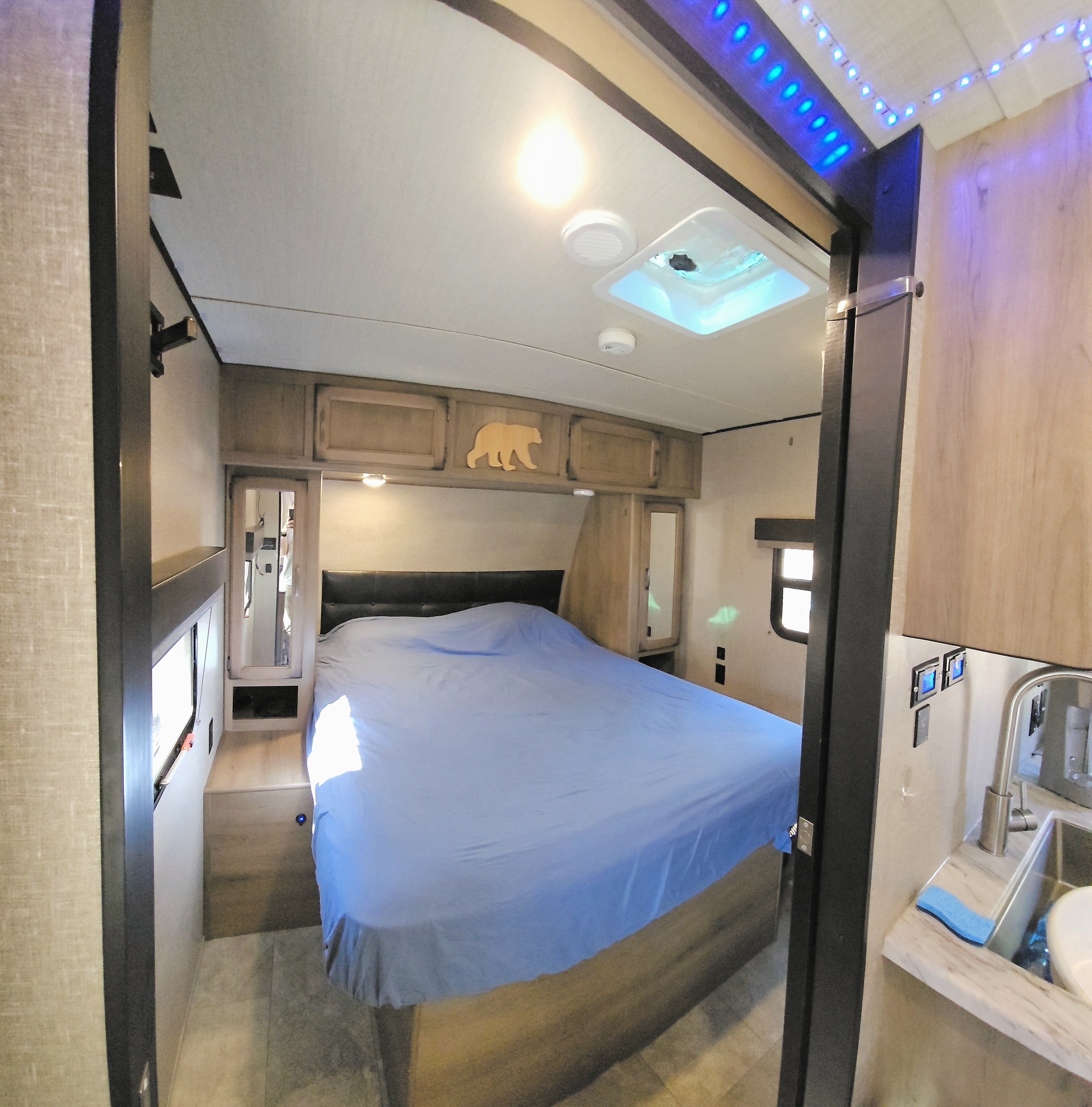 Inside an RV rental: a cozy bedroom with a blue bed, handy cabinets, and a small sink, perfect for mobile adventures.. Grand Design Transcend 2019