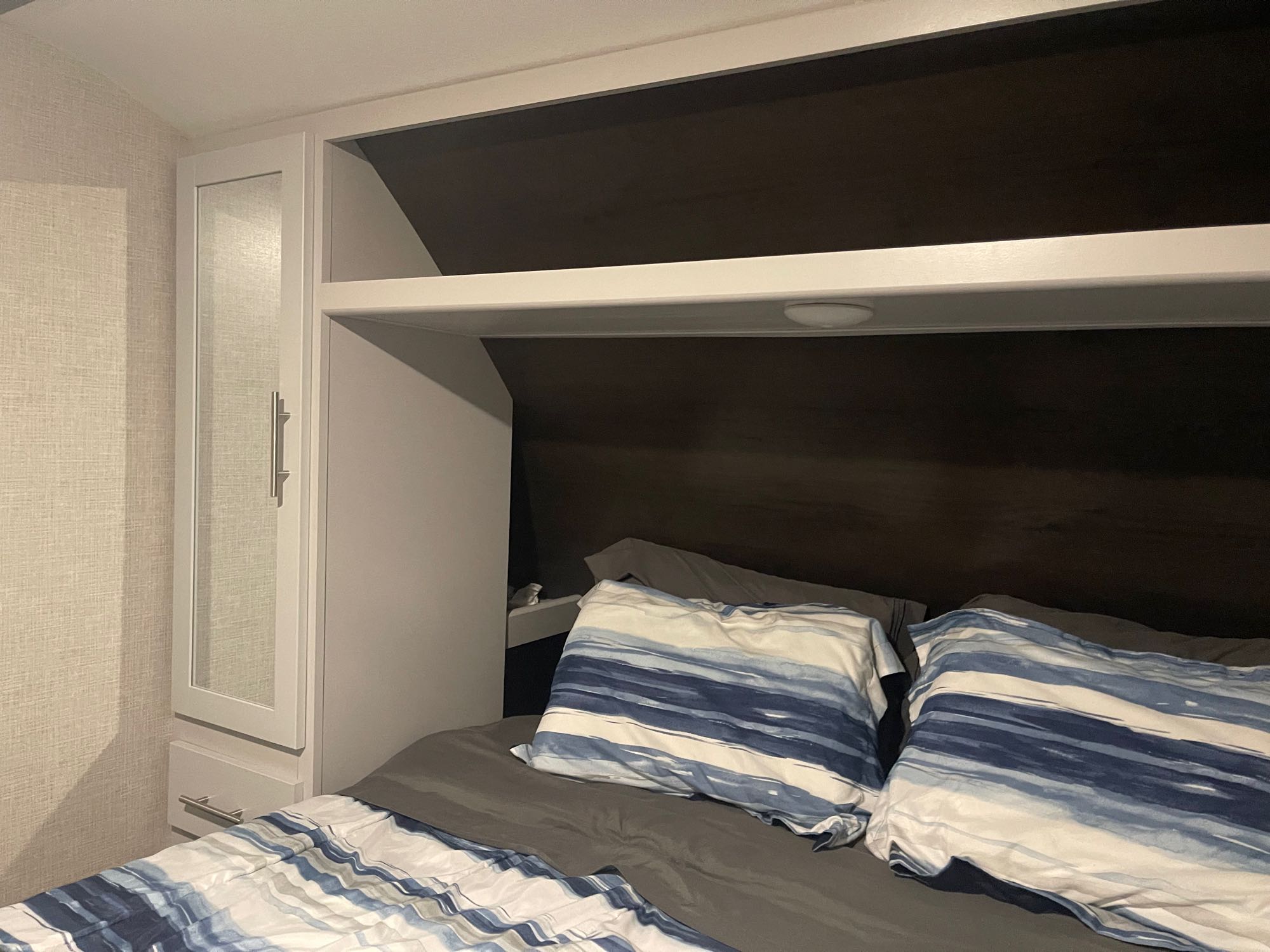 Compact RV rental bedroom featuring a built-in mirror cabinet, overhead storage, and blue striped bedding.. Keystone RV Bullet Ultra Lite 2021