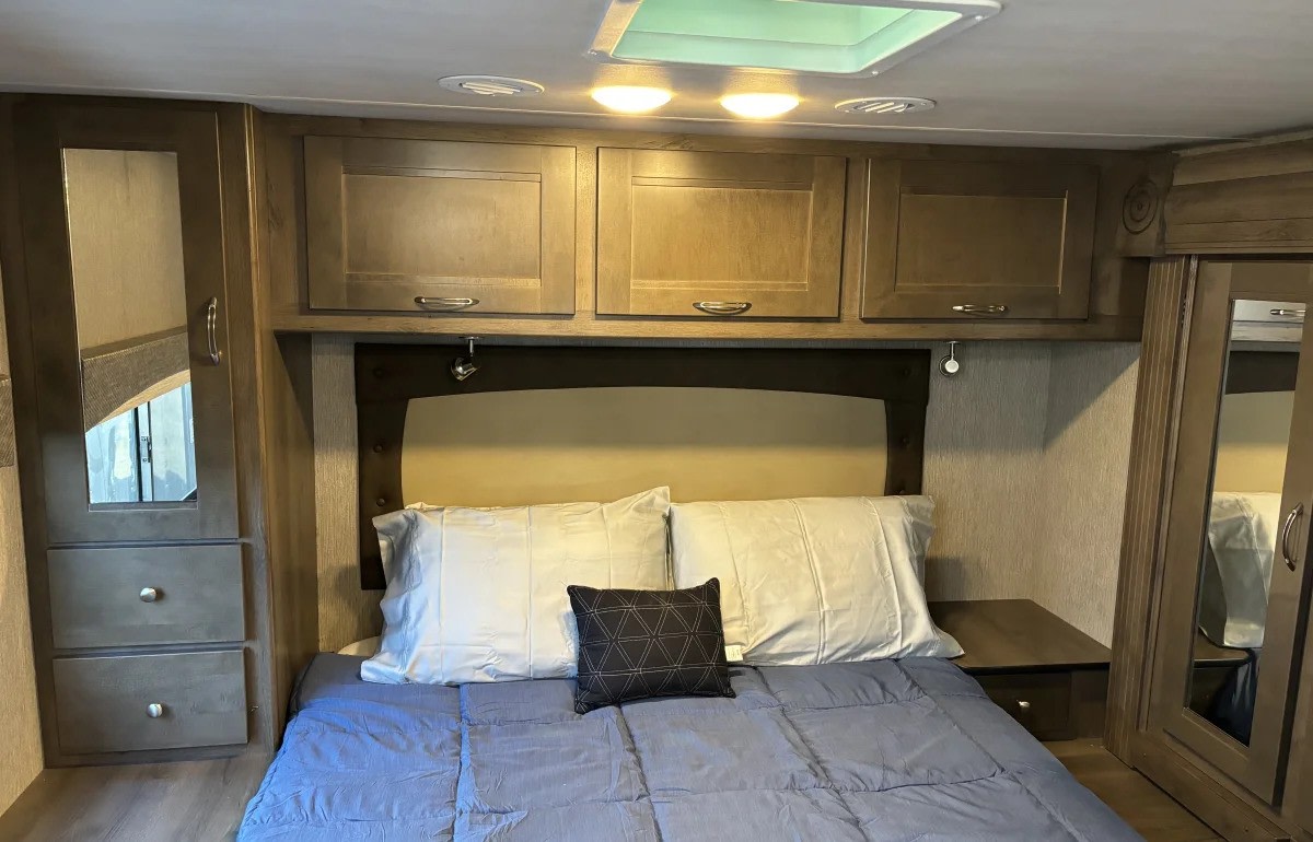 Compact RV rental bedroom showing a bed with blue comforter, overhead cabinets, and built-in storage on both sides.. Nexus RV Triumph 2021