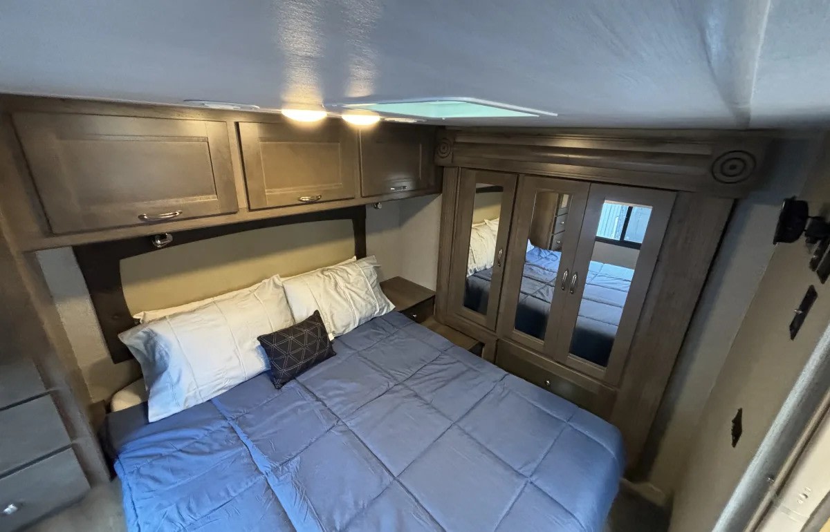 Inside a comfortable RV rental bedroom, featuring a bed, overhead cabinets, and mirrored closet. Ideal for travel.. Nexus RV Triumph 2021