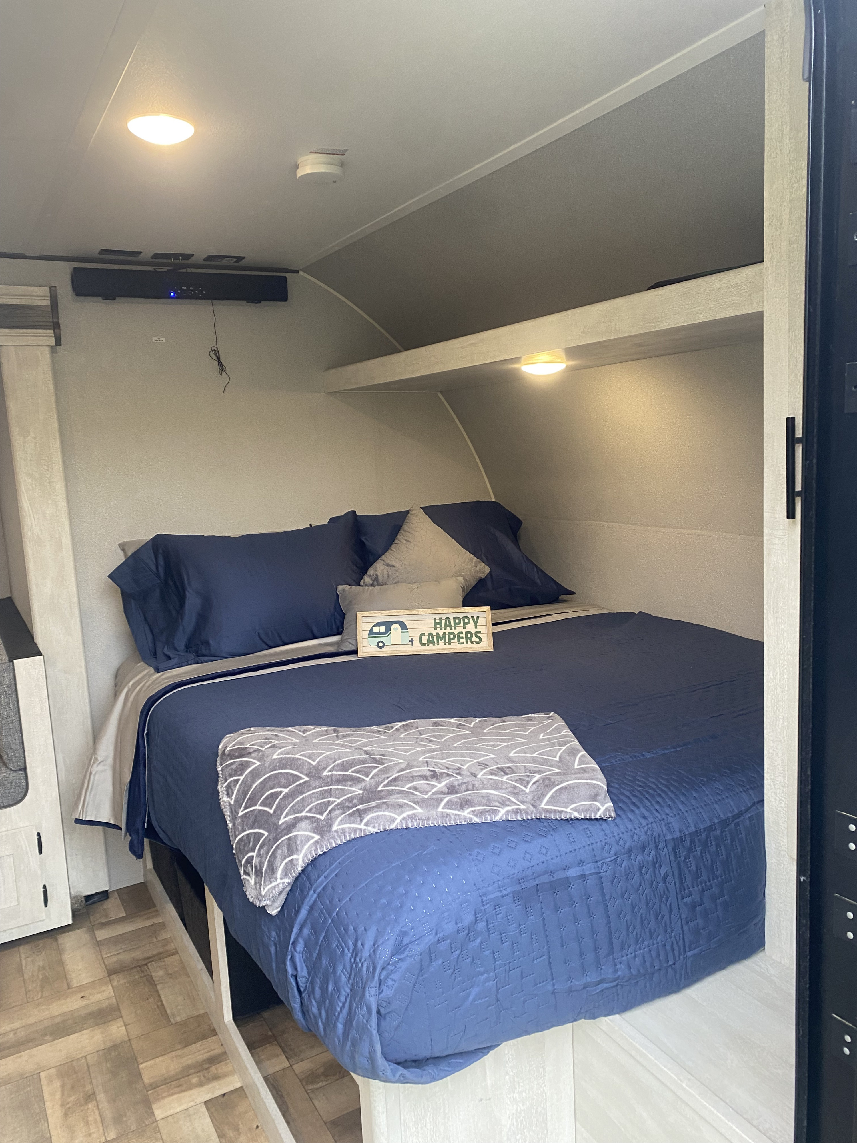 Cozy RV rental bedroom with a large bed, navy bedding, and 'Happy Campers' decor. Features built-in storage and sound system.. Forest River Shasta 2023