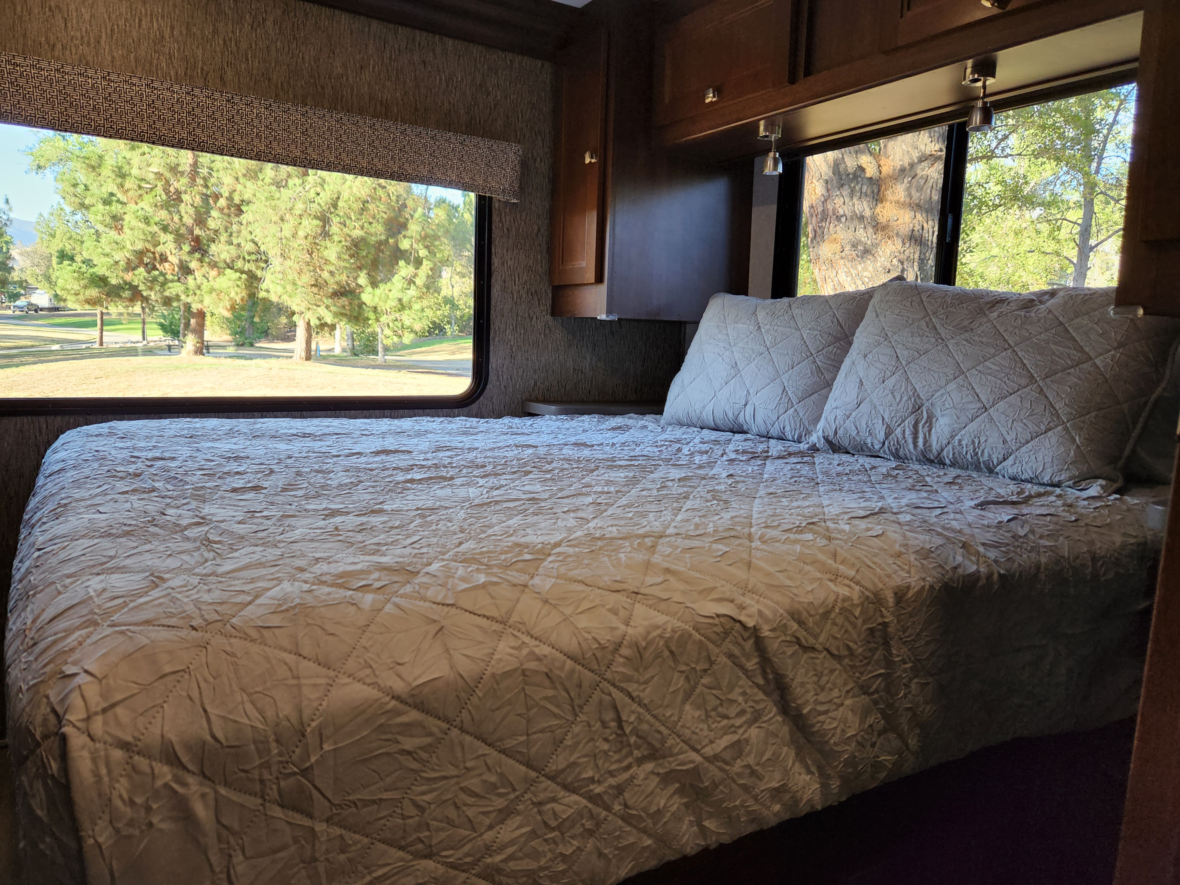 Cozy RV bedroom in an RV rental, featuring a quilted bed, reading lights, and a scenic park view.. Forest River Sunseeker 2018