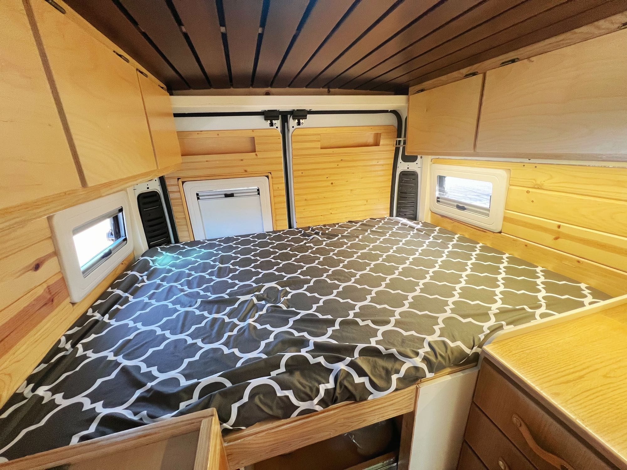 Comfortable camper van interior for RV rental, showcasing a patterned bed, light wood walls, ceiling, and storage.. Ram promaster 2500 2017