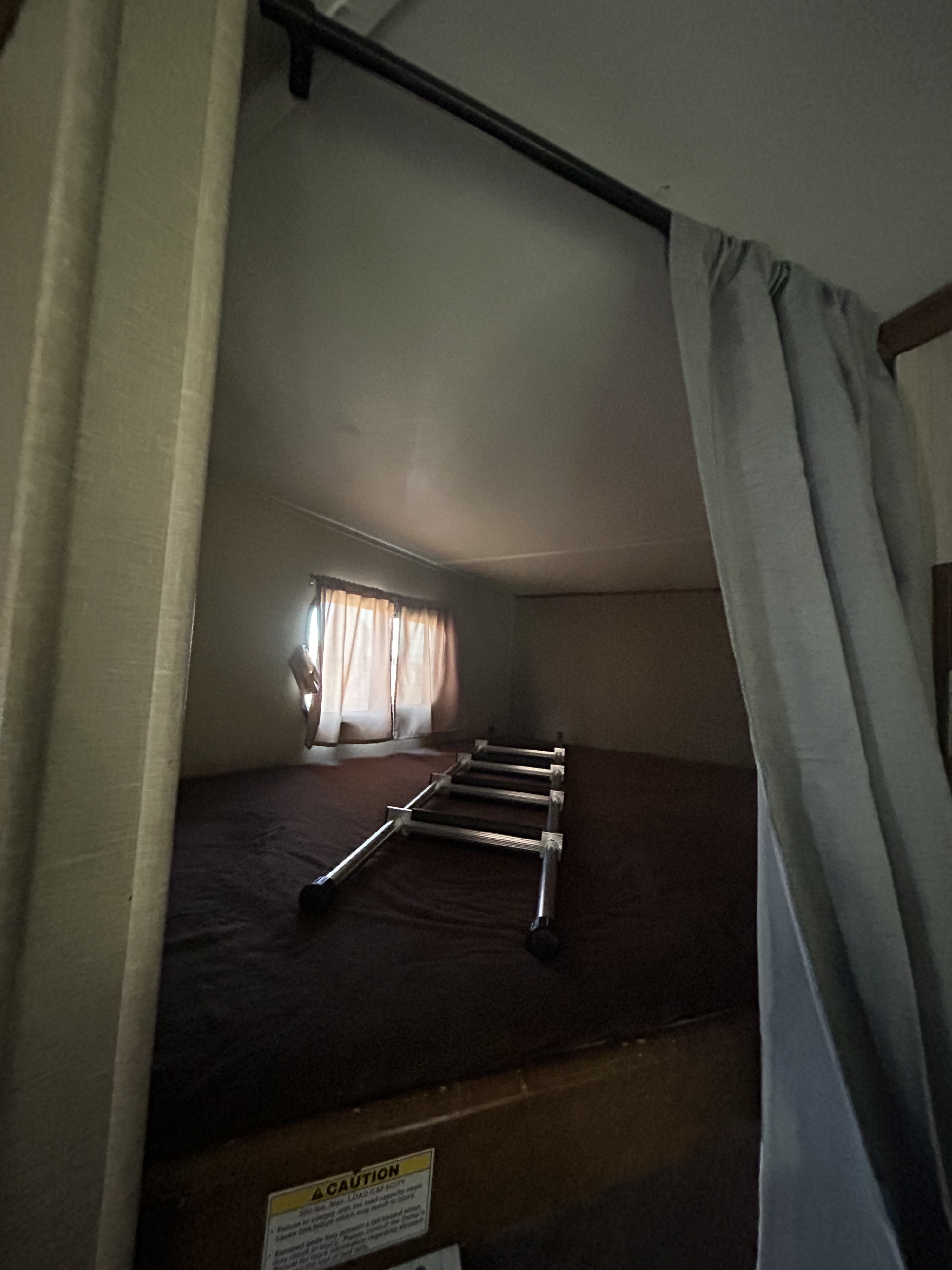 This RV rental features a compact overhead sleeping bunk with a ladder lying on the mattress and a window with light curtains.. Crossroads RV Rezerve 2016