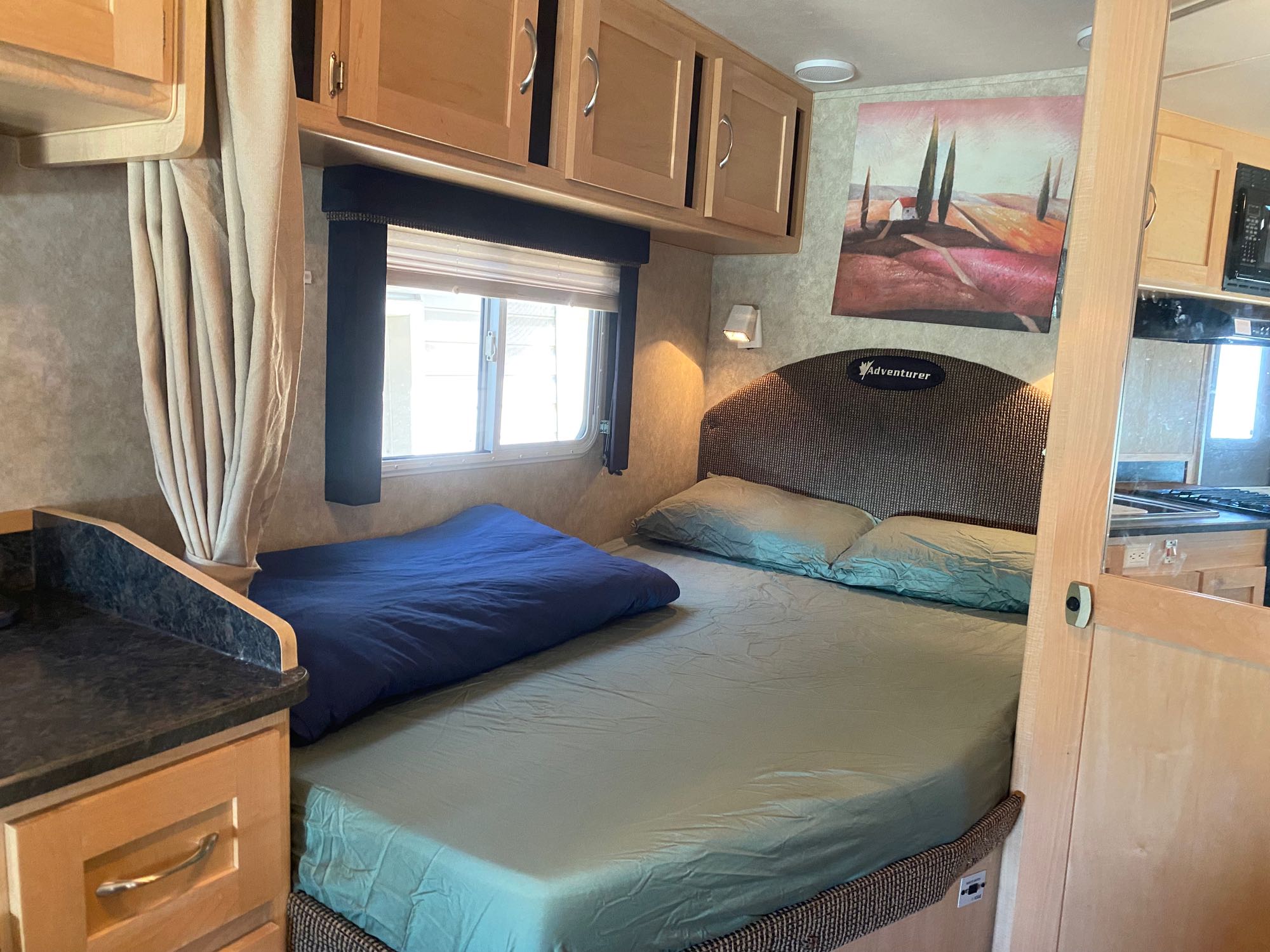 This RV rental bedroom features a comfy bed with green sheets, overhead cabinets, a window, and artistic landscape decor.. Adventurer Adventurer Motorhome 2010