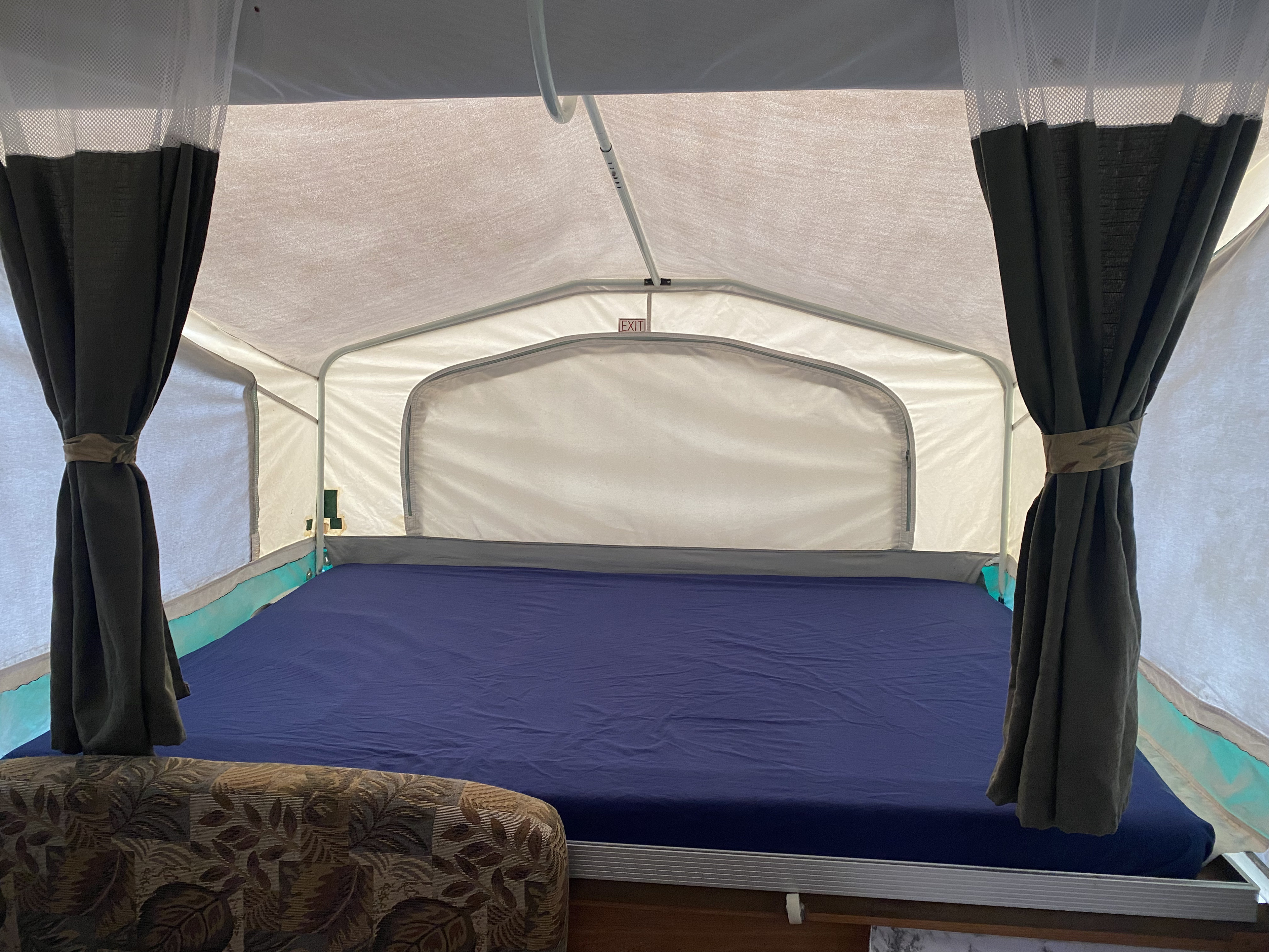 Inside this **RV rental** pop-up camper, a neat bed with blue sheets and tied dark curtains offers a cozy retreat. An "EXIT" sign is visible.. Palomino Real - Lite 801 2007