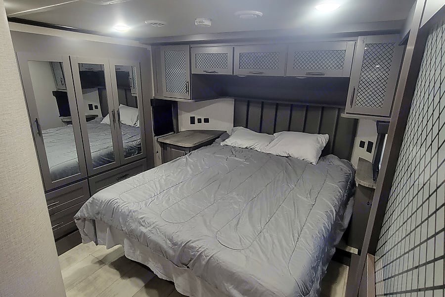 Modern RV rental bedroom with a large bed, stylish grey cabinets, mirrored wardrobes, and contemporary decor for your travels.. Grand Design Momentum M-Class Toy Hauler 2022