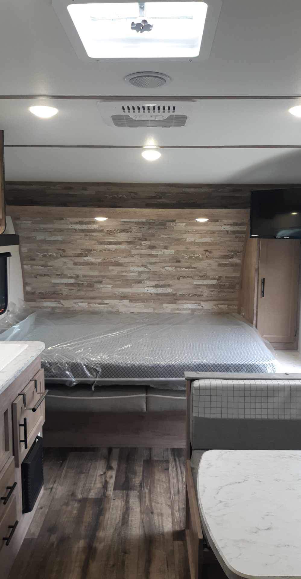 RV rental interior: Bedroom with plastic-wrapped bed, wooden accent wall, TV, and adjacent dining booth. Features ceiling AC.. Grand River 22MB 2024
