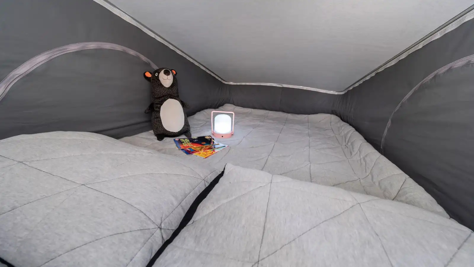 Cozy RV rental pop-top tent interior, featuring a soft bed, stuffed bear, lit lamp, and books for a comfortable adventure.. Winnebago 59P 2023