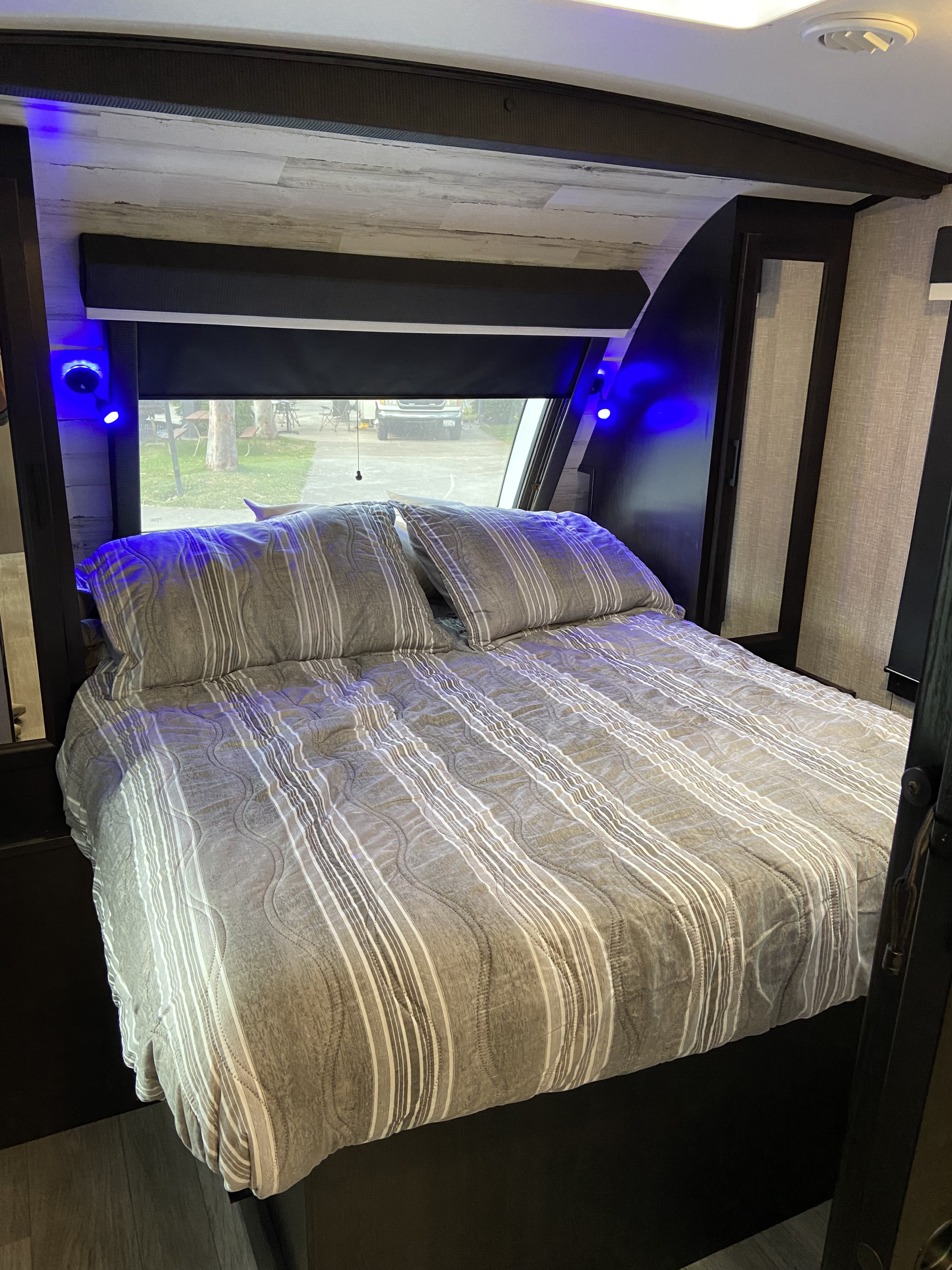 Cozy RV rental bedroom featuring striped bedding, blue accent lights, and a view of the campsite outside. Ideal for travel accommodations.. Jayco White Hawk 2023