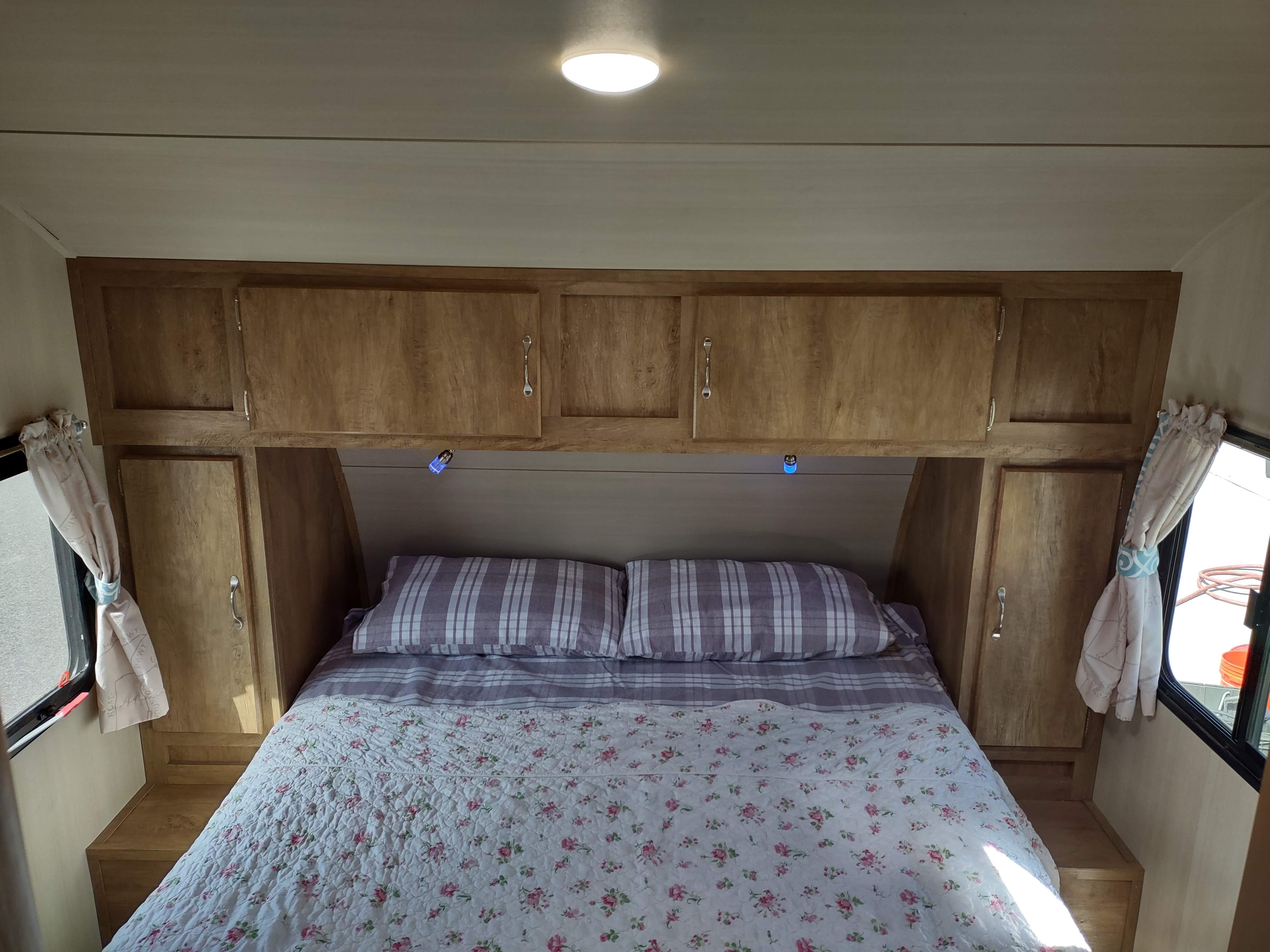 This RV rental offers a cozy bedroom with a floral quilt, plaid pillows, and wood-toned overhead cabinets for storage.. Gulf Stream Vintage Cruiser 2020