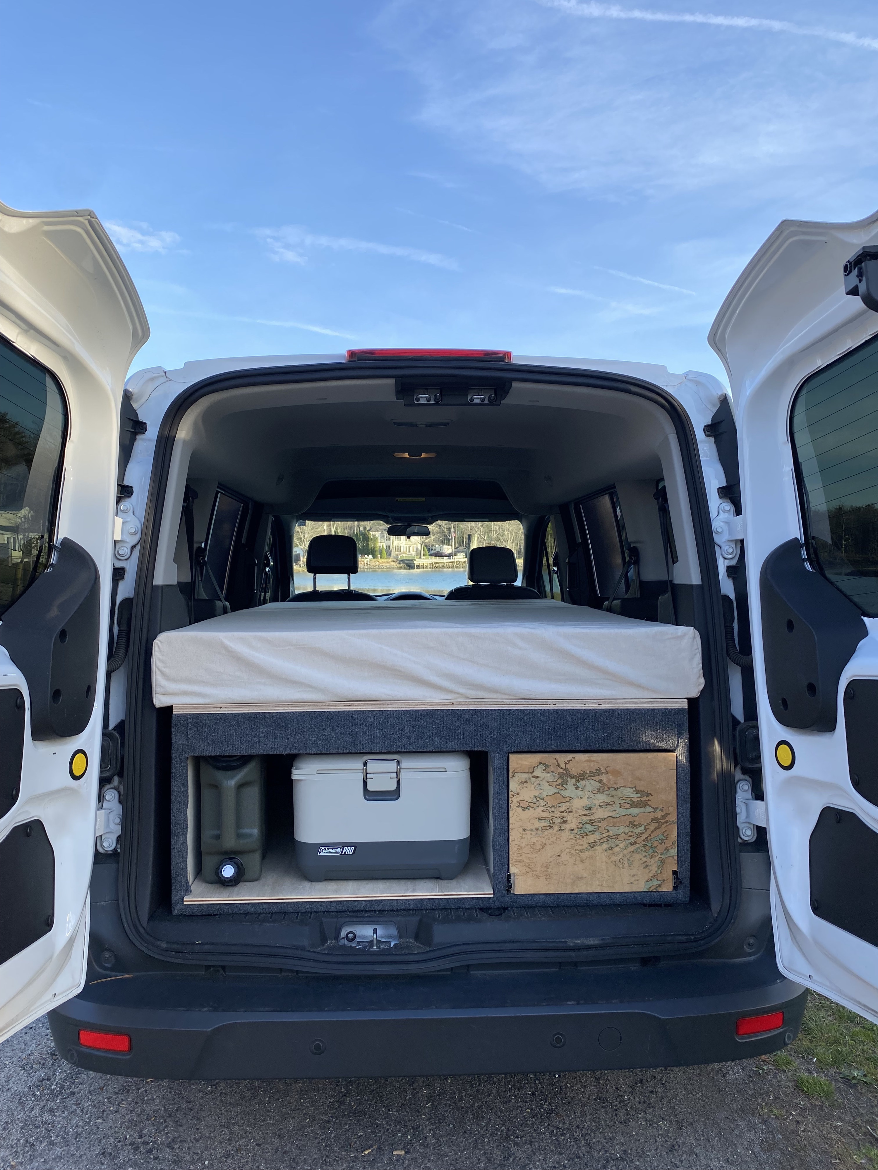 Rear view of a white camper van's interior, featuring a bed, cooler, water jug, and storage. Ideal for an rv rental adventure by the water.. Ford Transit Connect 2017