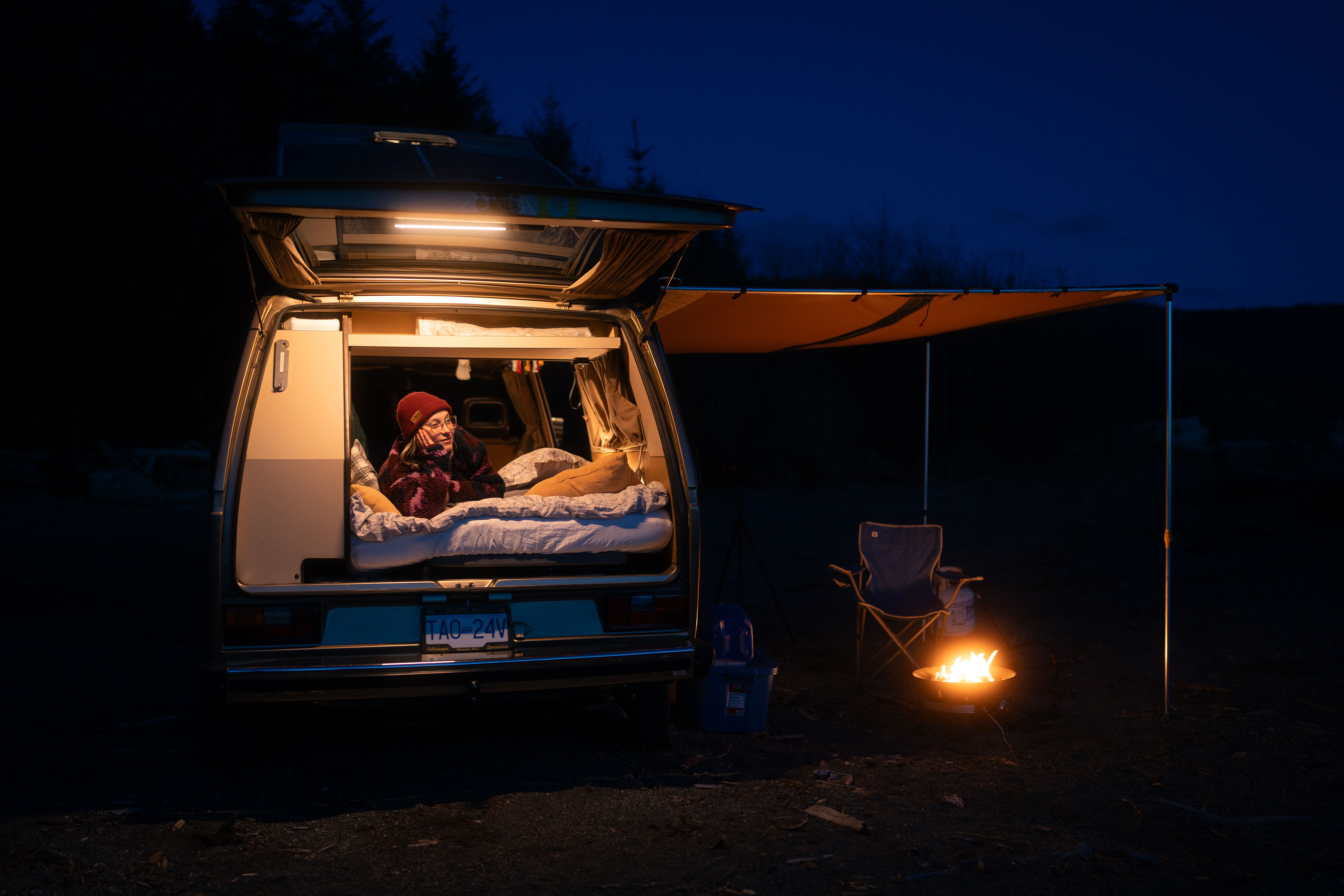 Enjoy van life with an RV rental! A person relaxes in a cozy, lit camper van at night. A warm fire and awning await outside.. Volkswagen Westfalia 1989