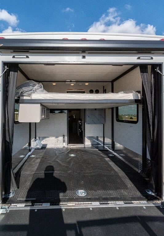 Inside an RV rental toy hauler: A spacious rear garage with a high-mounted bed, durable black floor, and ramp door open to the sky.. Thor Outlaw 2024