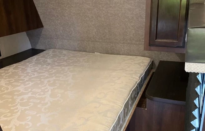 Inside an RV rental: a compact bedroom features a patterned mattress on a bed base, dark wood cabinetry, and textured walls.. Jayco Jay Flight SLX 2019
