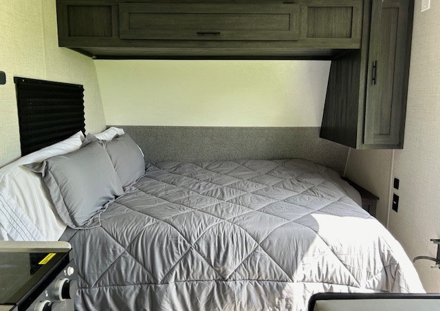 Cozy RV rental bedroom showcasing a grey quilted bed, overhead cabinets, and a window for comfortable travel.. Forest River Ozark 2021
