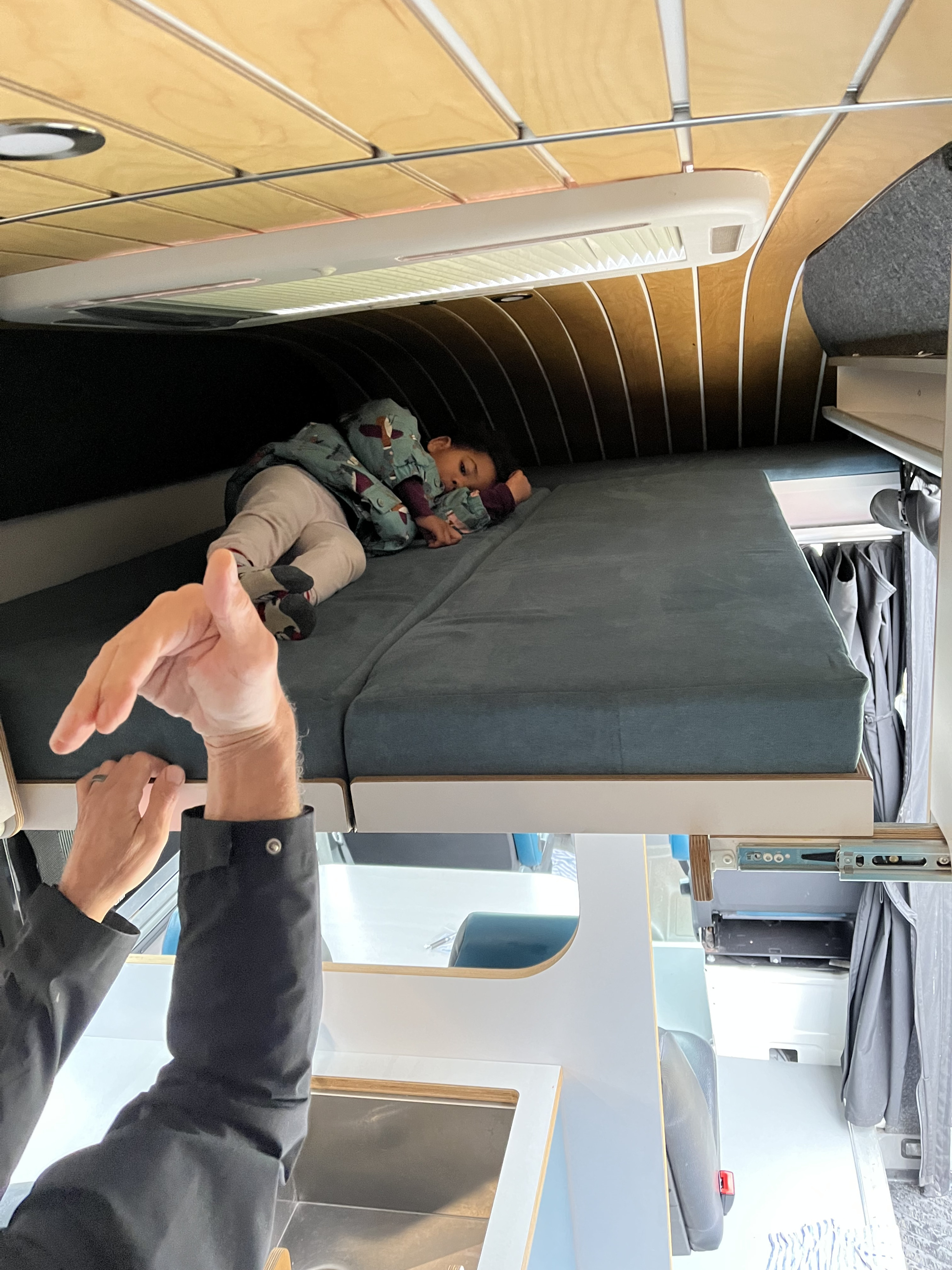 Inside a custom RV rental: A child naps peacefully on a comfortable bed with wooden ceiling details & kitchen below.. Mercedes Sprinter 2015