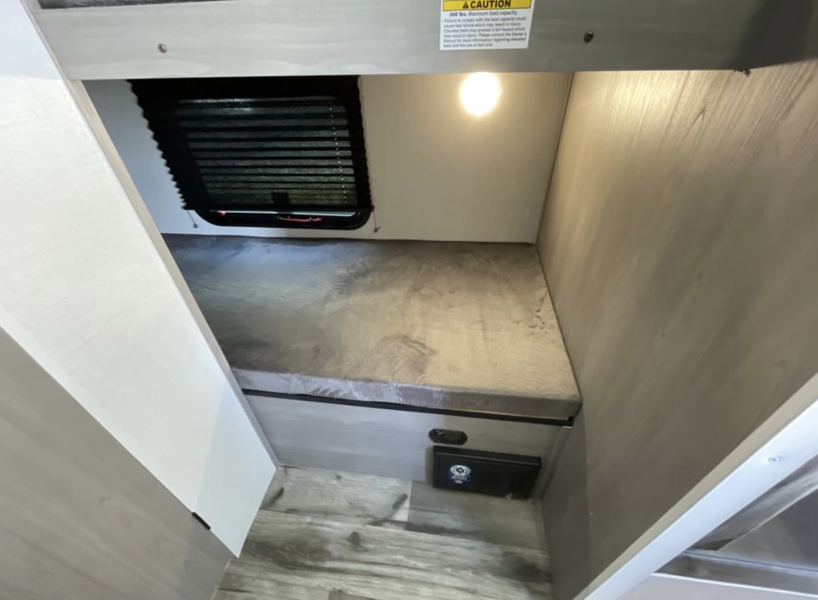Compact RV rental lower bunk features a cozy gray mattress, window with blinds, overhead light, and unit controls below.. Coleman Lantern 17B 2024