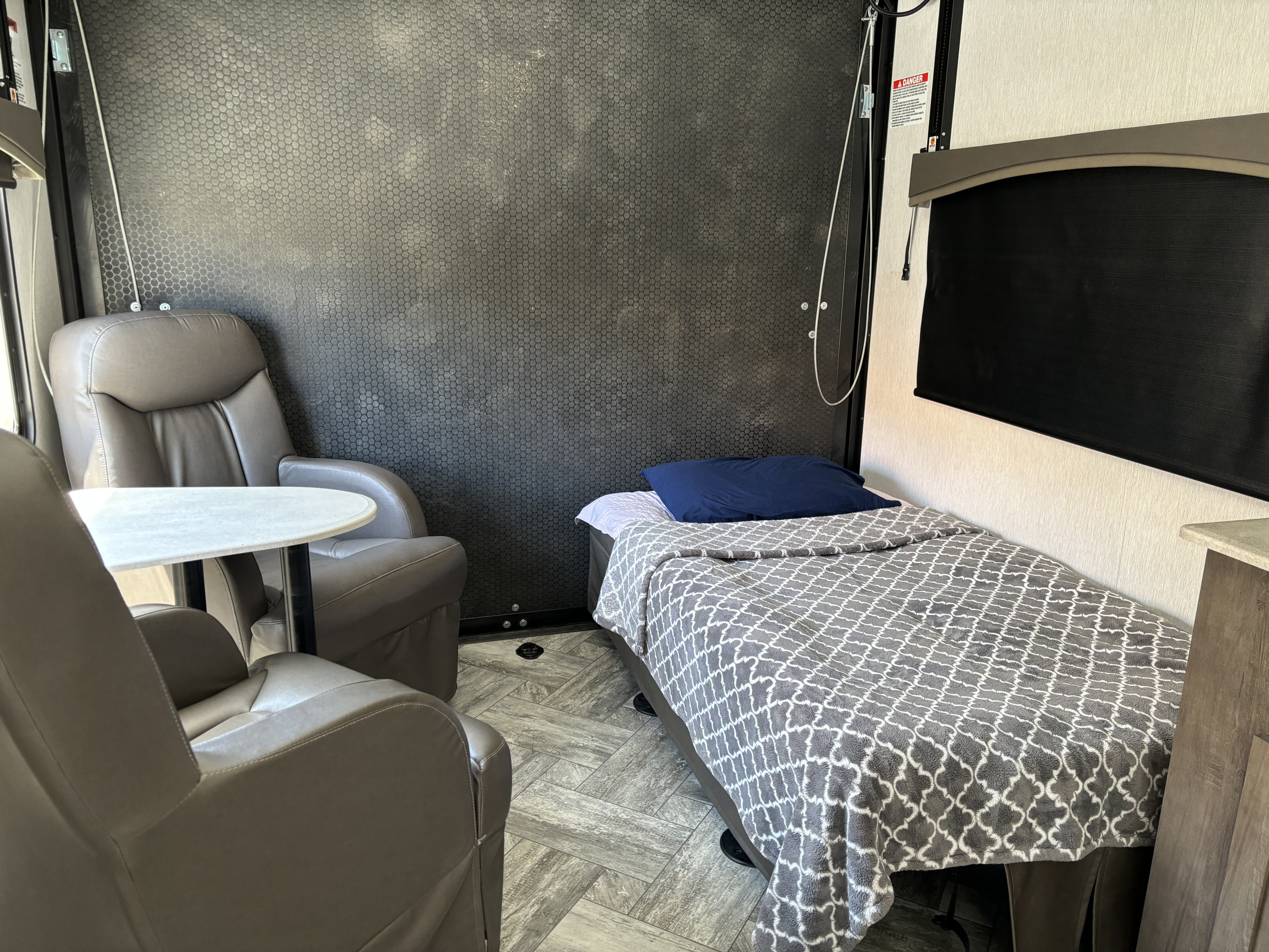 Inside an RV rental: two gray recliners, a small table, and a bed with a patterned blanket.. Forest River Stealth 2022