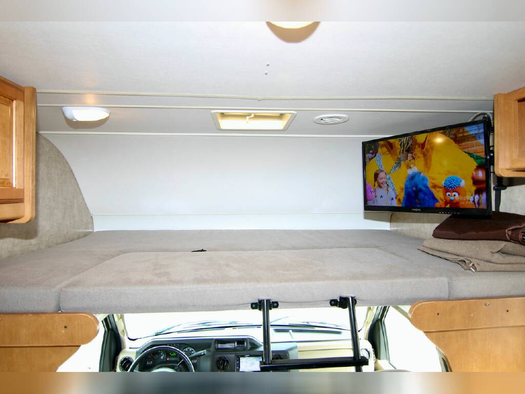 RV rental interior: comfy over-cab bunk bed and a mounted TV playing a kids' show, ideal for families.. Winnebago Minnie Winnie 2016