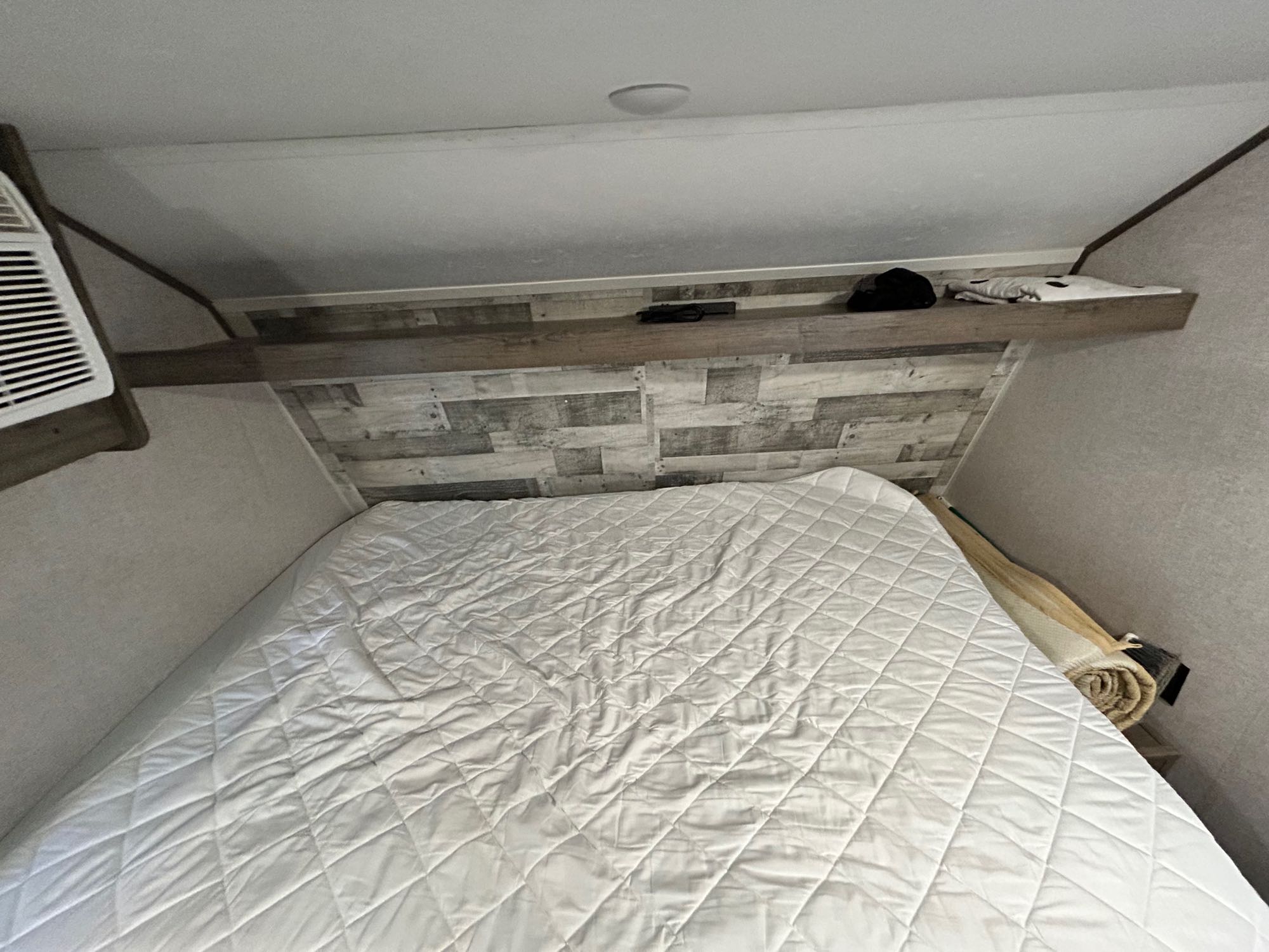 Compact RV rental sleeping area featuring a white quilted bed, wood accent wall, overhead shelf, and AC unit. Ready for adventure.. Aspen Trail LE 2021