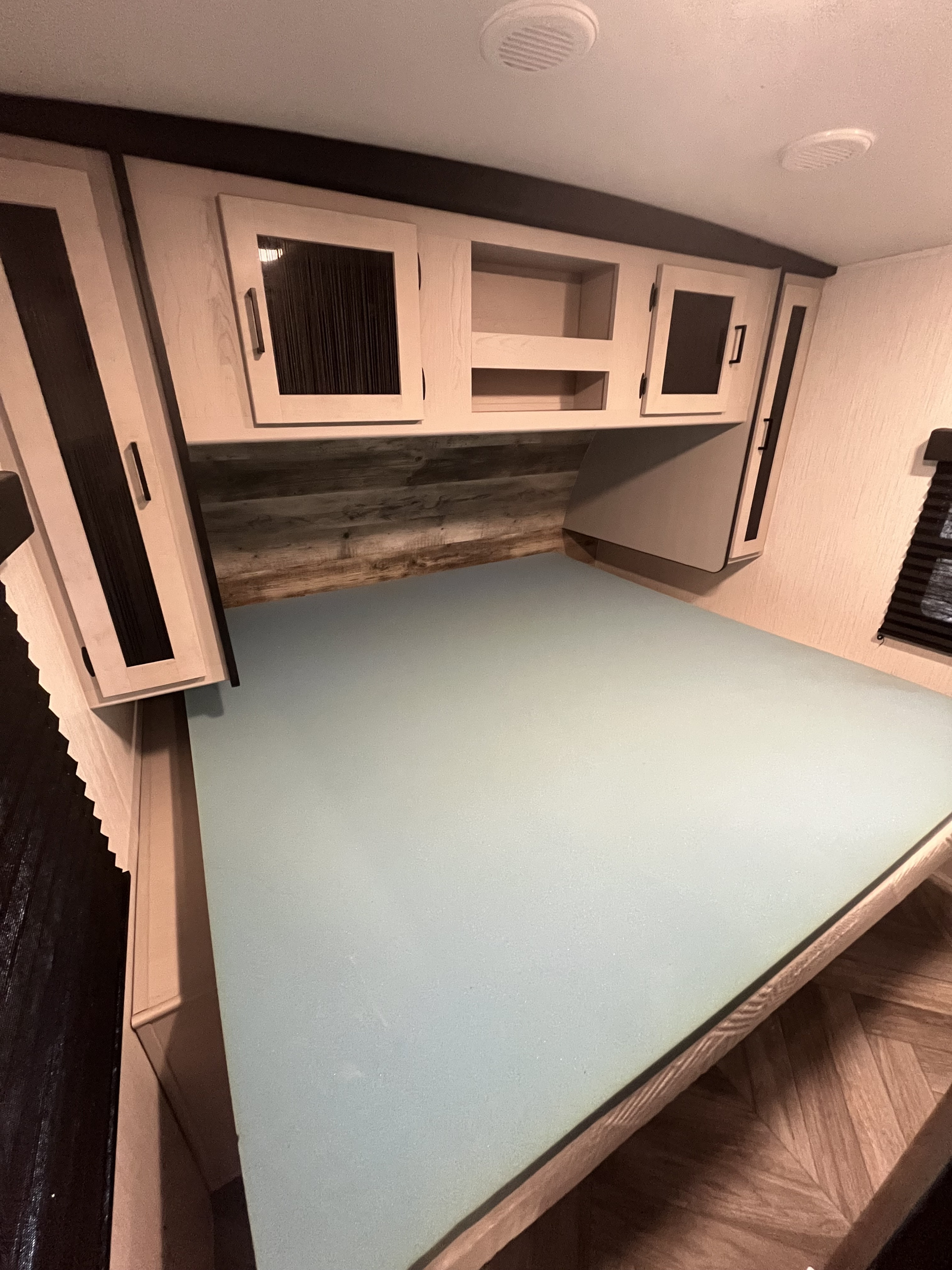 Compact RV rental bedroom featuring a light blue-green mattress, overhead cabinets, and wood-plank headboard.. Heartland Mallard 2023