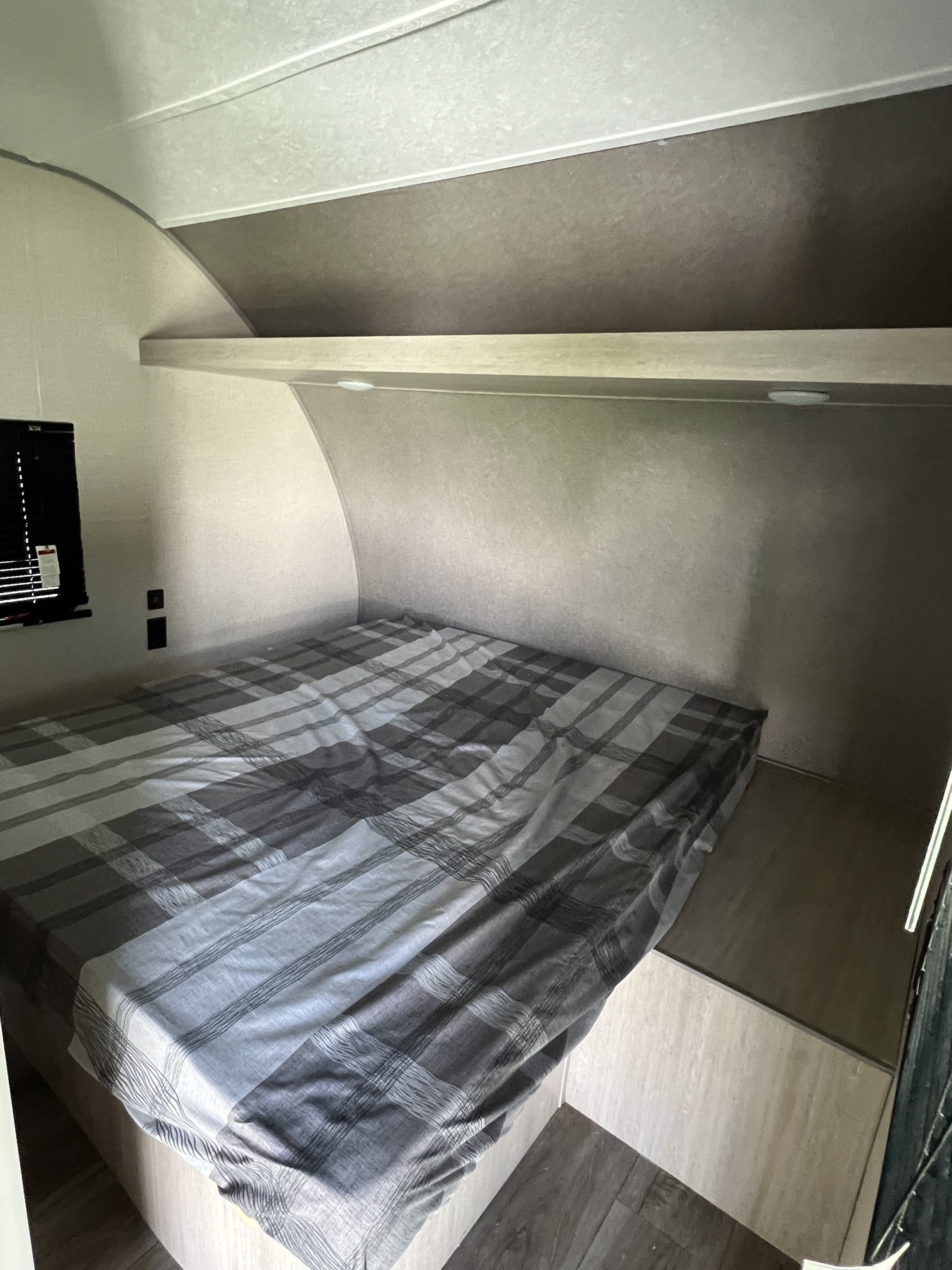RV rental bedroom interior with a grey plaid bed, overhead shelf with lights, and compact curved walls.. Coachmen Catalina 2022