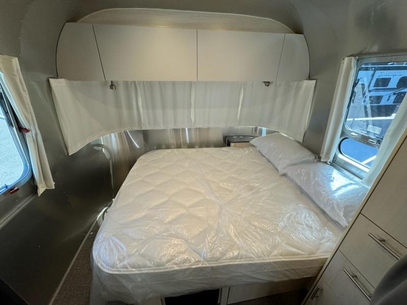 Inside an RV rental unit: a bright bedroom with a bed & pillows covered in plastic, overhead storage, and windows.. Airstream Flying Cloud 2023