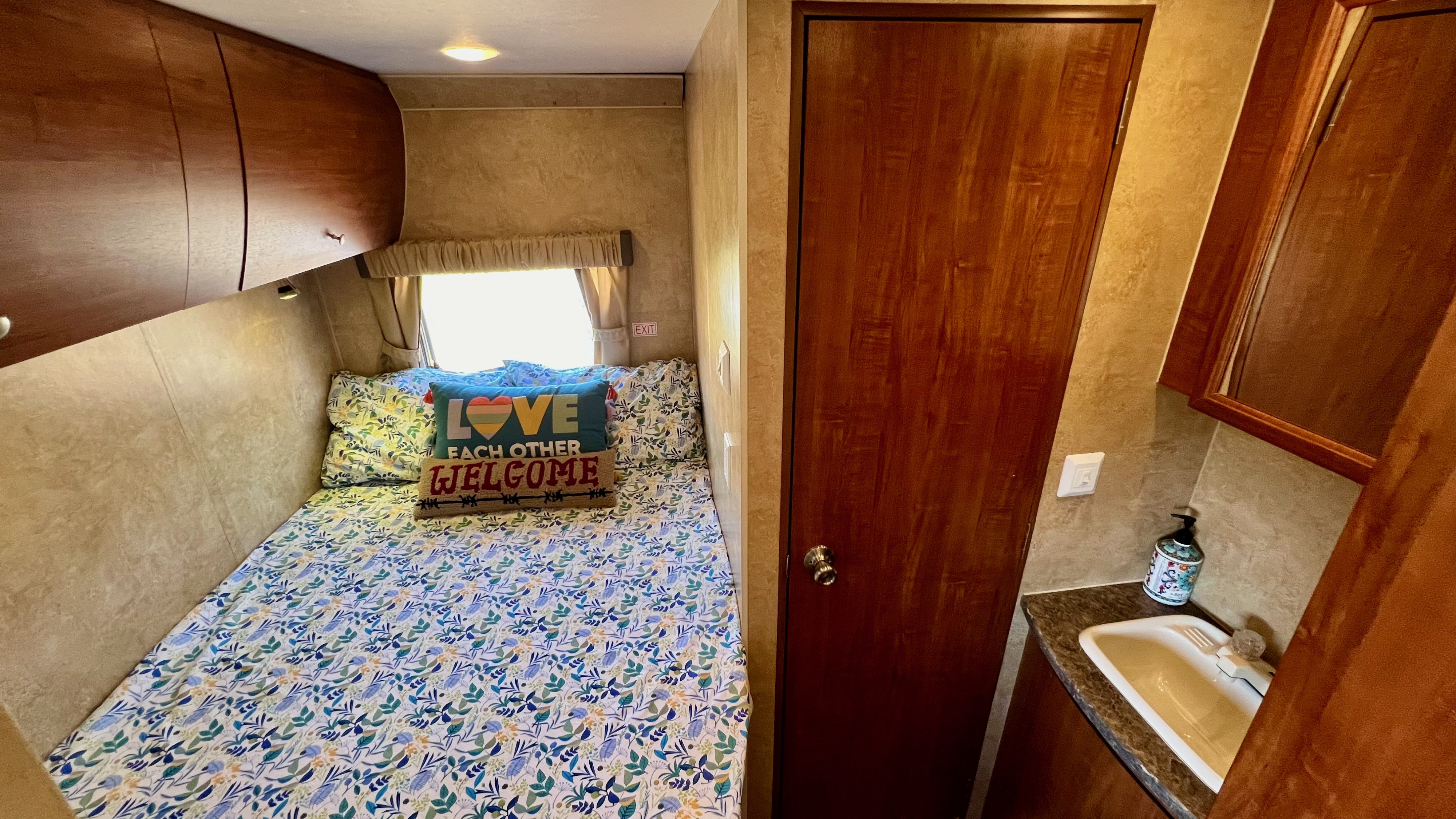 RV rental interior: cozy bedroom with leaf-patterned bed, "LOVE" pillow, overhead cabinets, and a compact sink area.. Jayco Skylark 2011