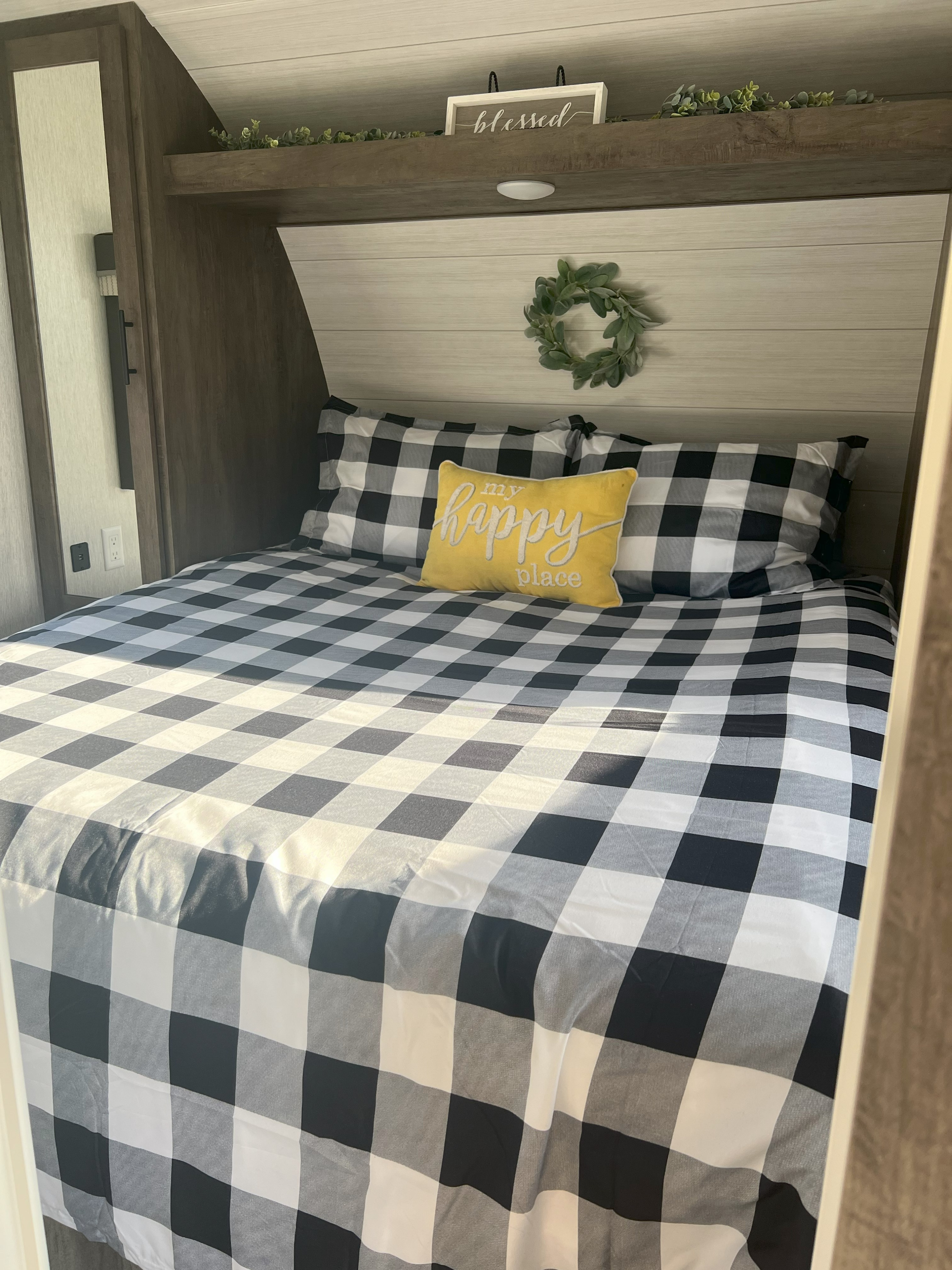 Cozy RV rental bedroom with black and white buffalo plaid bedding, a yellow 'my happy place' pillow, and decorative wreath.. Forest River Wildwood X-Lite 2022