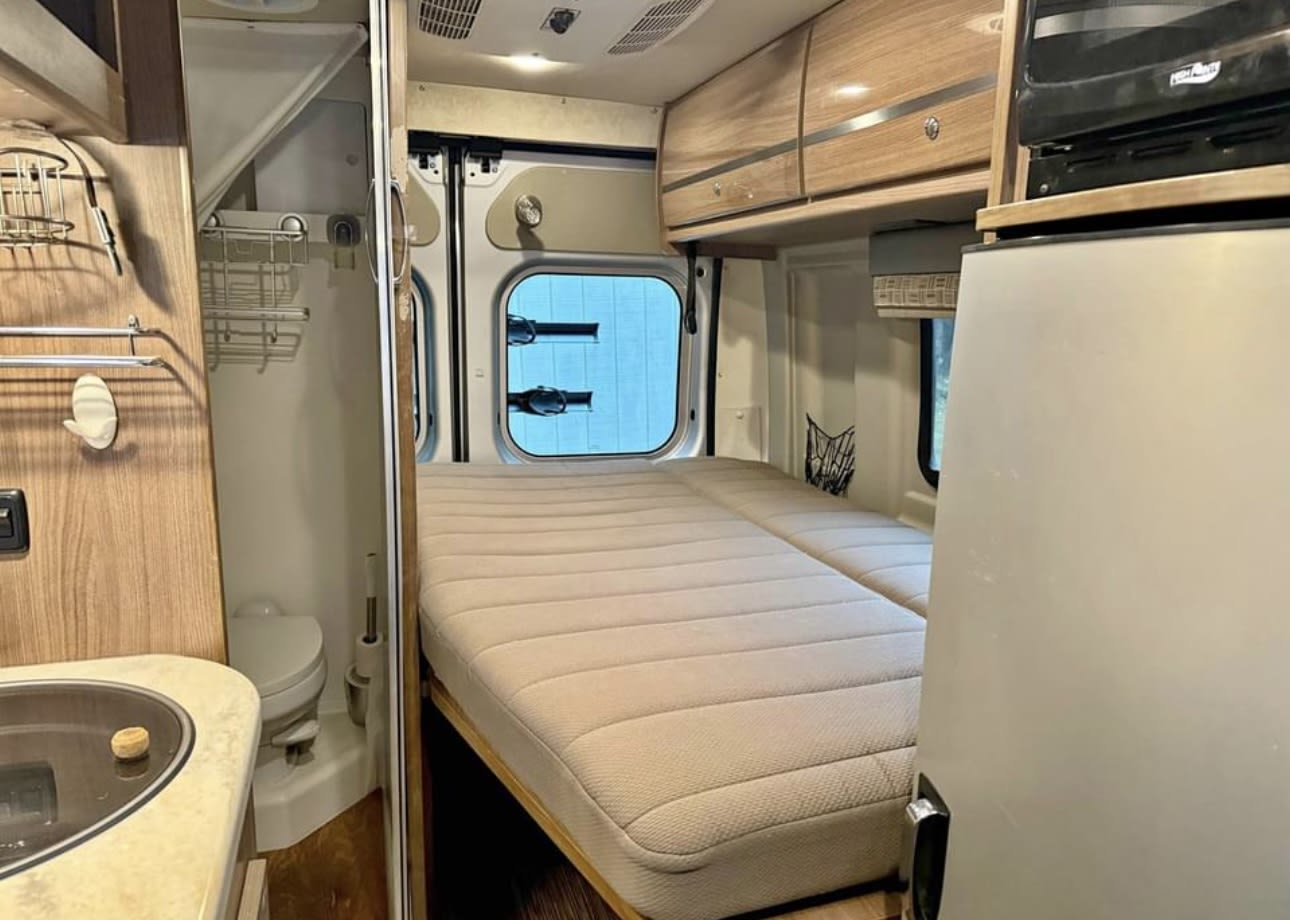 View inside an RV rental: a plush bed, compact bathroom, and kitchenette with sink, fridge, and microwave.. Winnebago Travato 2015