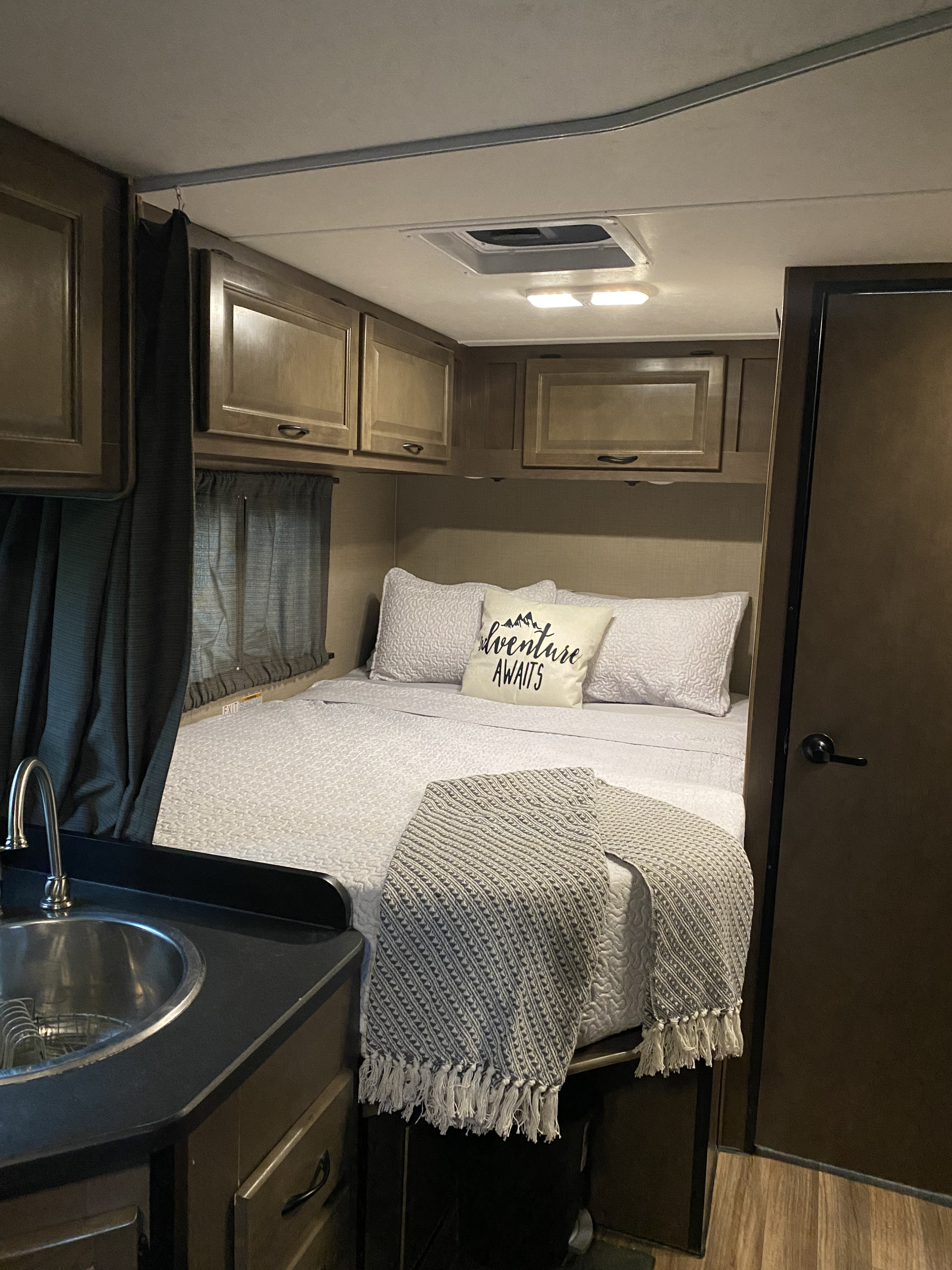 Cozy RV rental interior featuring a comfortable bed with an "Adventure Awaits" pillow and a compact kitchenette.. Thor Majestic 2019