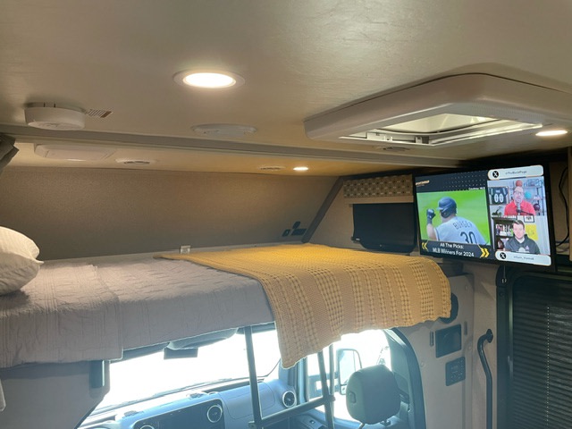 This RV rental interior features a comfortable bunk bed with a yellow blanket and a TV showing sports, ideal for travel.. Forest River Dynamax Isata 3 24FW 2023