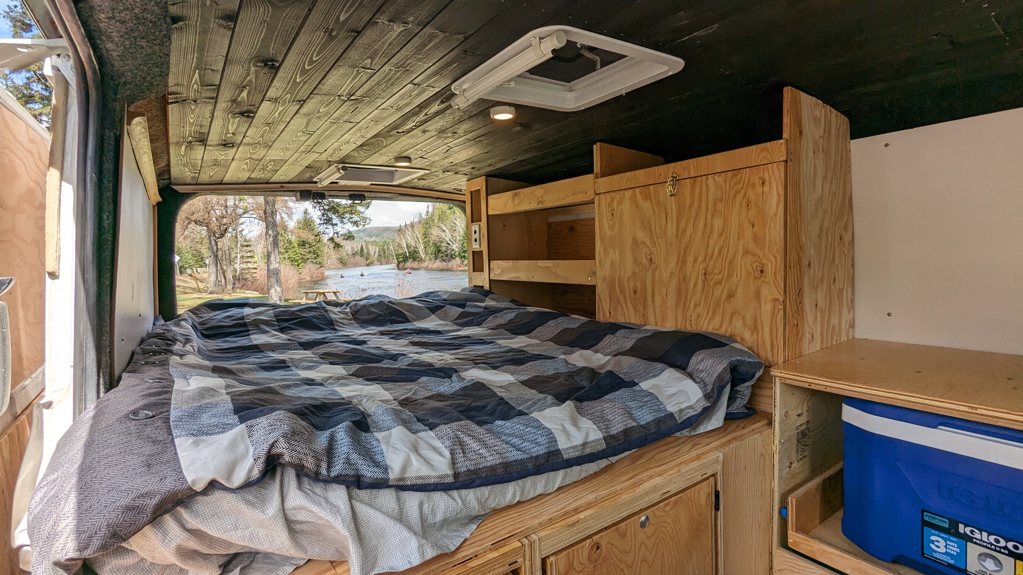RV rental camper van interior: cozy bed, rustic wood, and a scenic river view. Built-in storage and cooler shown.. Nissan Other 2012