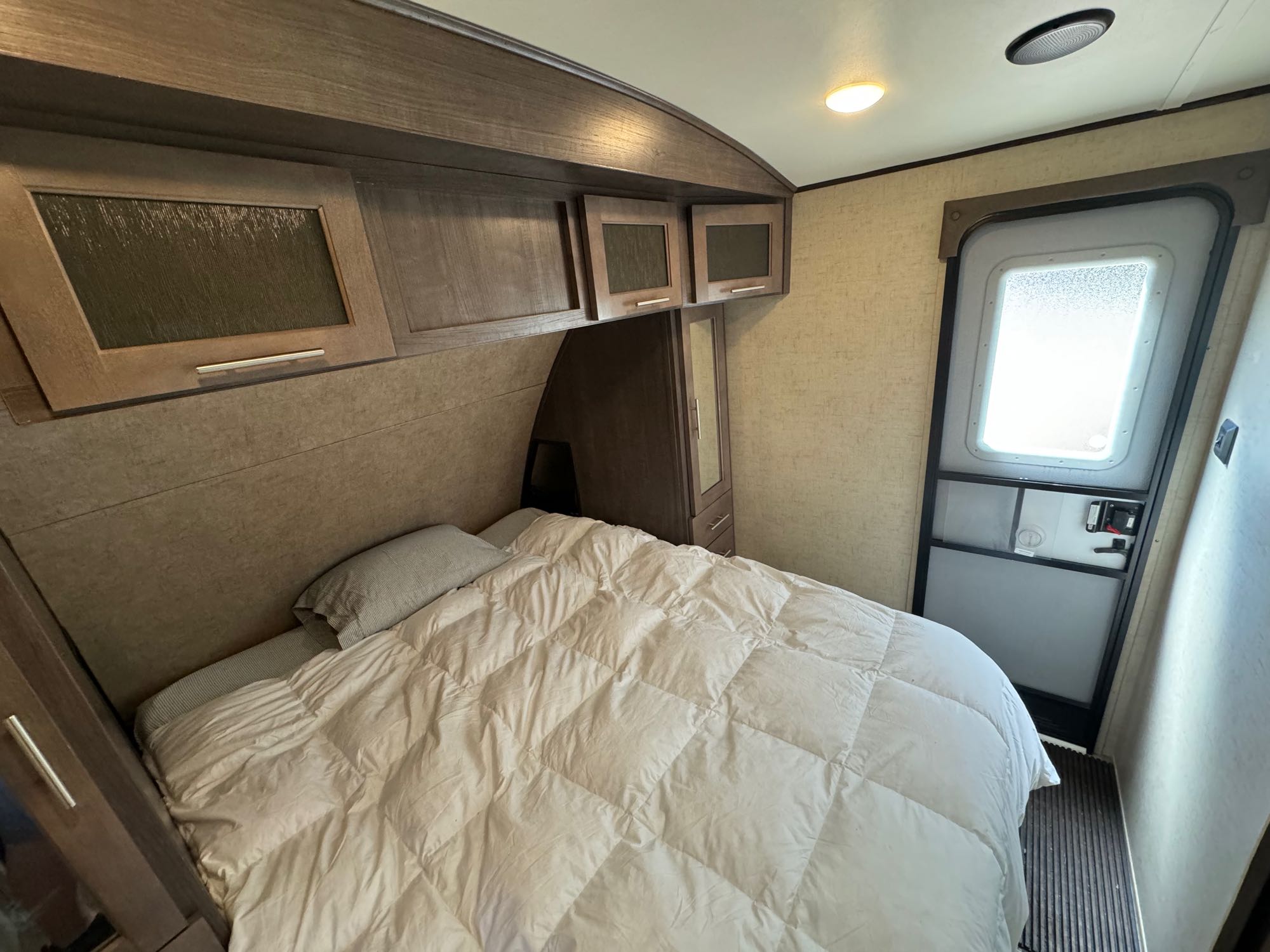 Cozy RV rental bedroom featuring a comfy bed with a white comforter, overhead storage cabinets, and an entry door.. Forest River Surveyor 2015