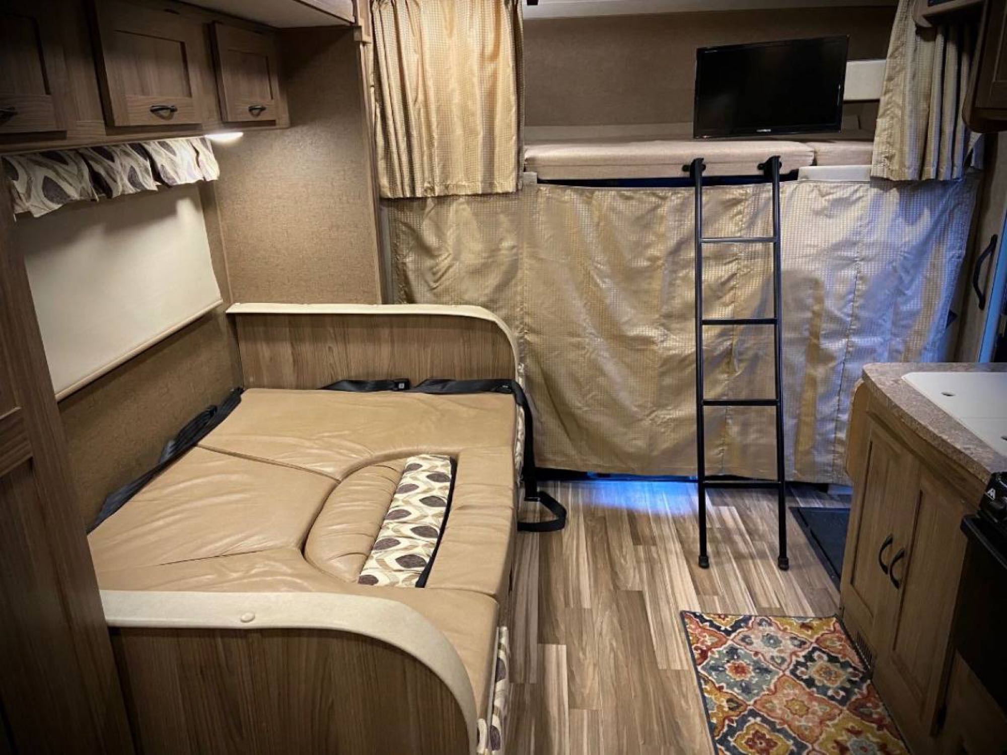 Cozy RV rental interior with convertible dinette, bunk beds, ladder, TV, and partial kitchen. Wood-look floors add warmth.. Coachmen Prism 2016