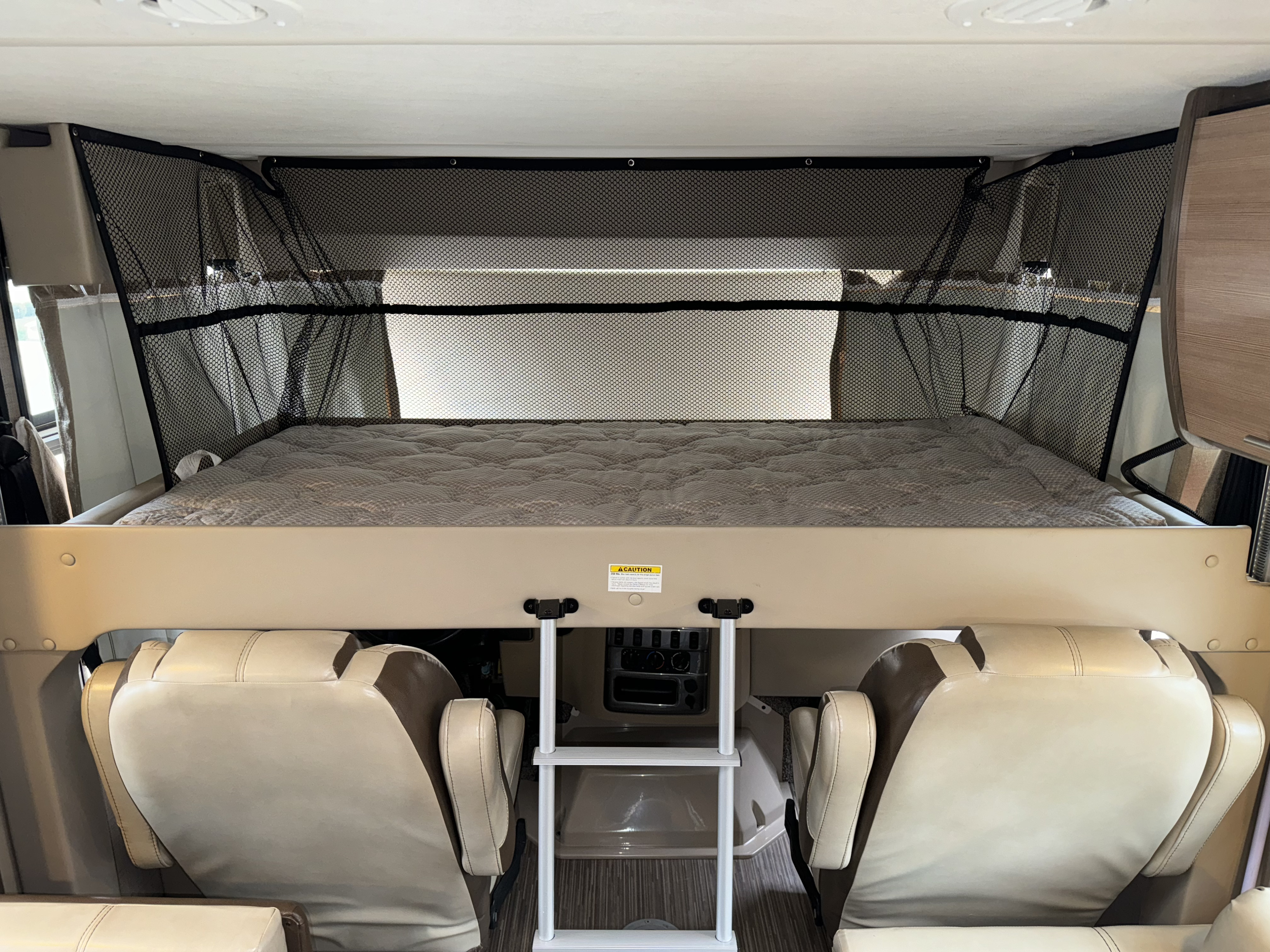 Inside an RV rental: overhead bunk bed with safety mesh, mattress, & ladder for access. Driver/passenger swivel seats below.. Thor Vegas 2018