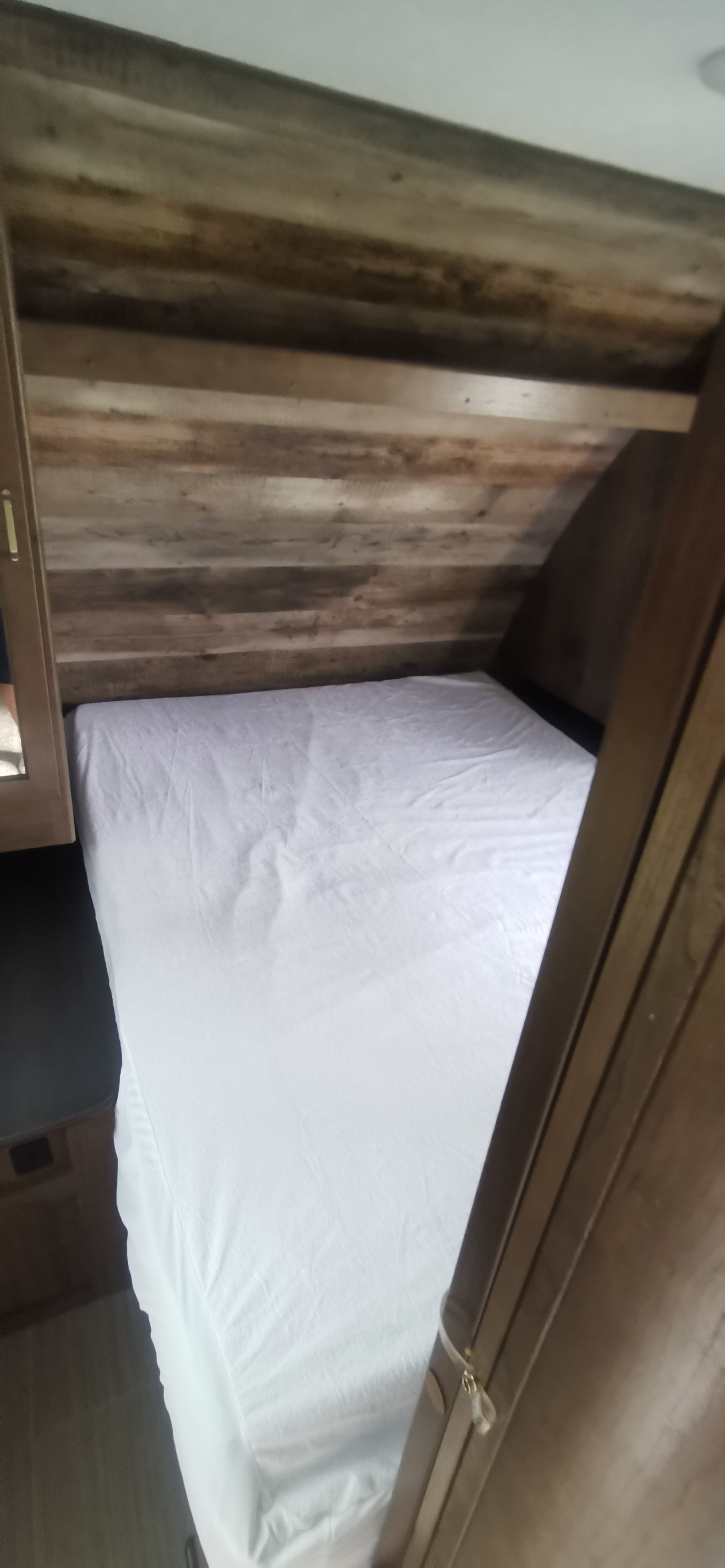 Cozy sleeping area in an RV rental. A bed with a white fitted sheet is tucked into a compact space with rustic wood-paneled walls.. Keystone RV Passport 2019