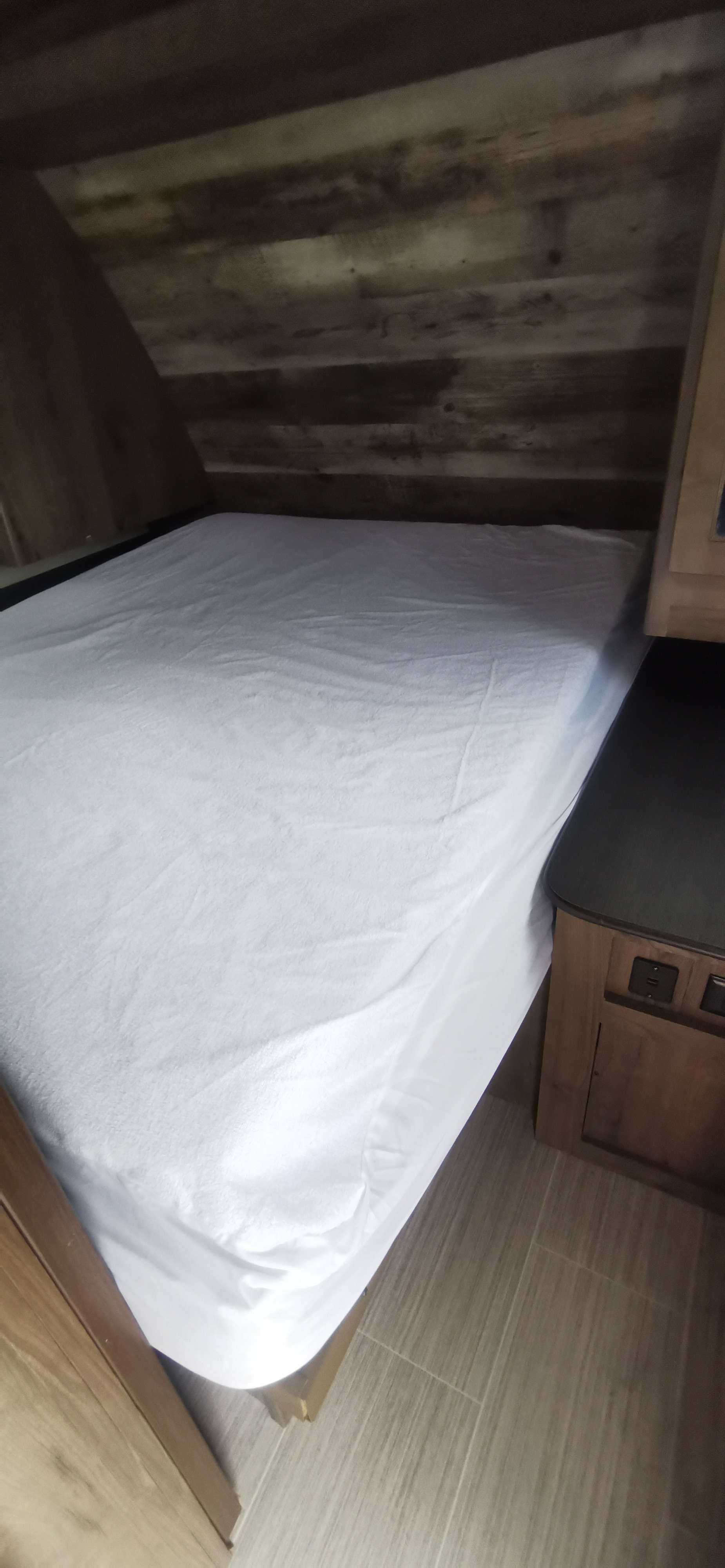 Cozy bed with white sheets in an RV rental's bedroom. Features rustic wood paneling, angled ceiling, and bedside charging outlets.. Keystone RV Passport 2019