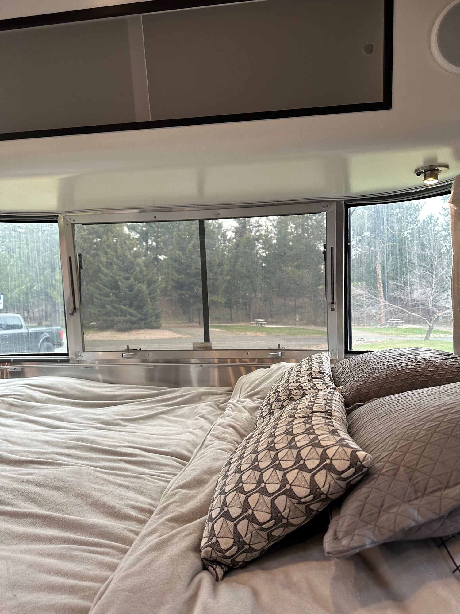 Inside an RV rental: a comfy bed with pillows facing large windows showing pine trees, a truck, and a park setting.. Airstream Caravelle 2020