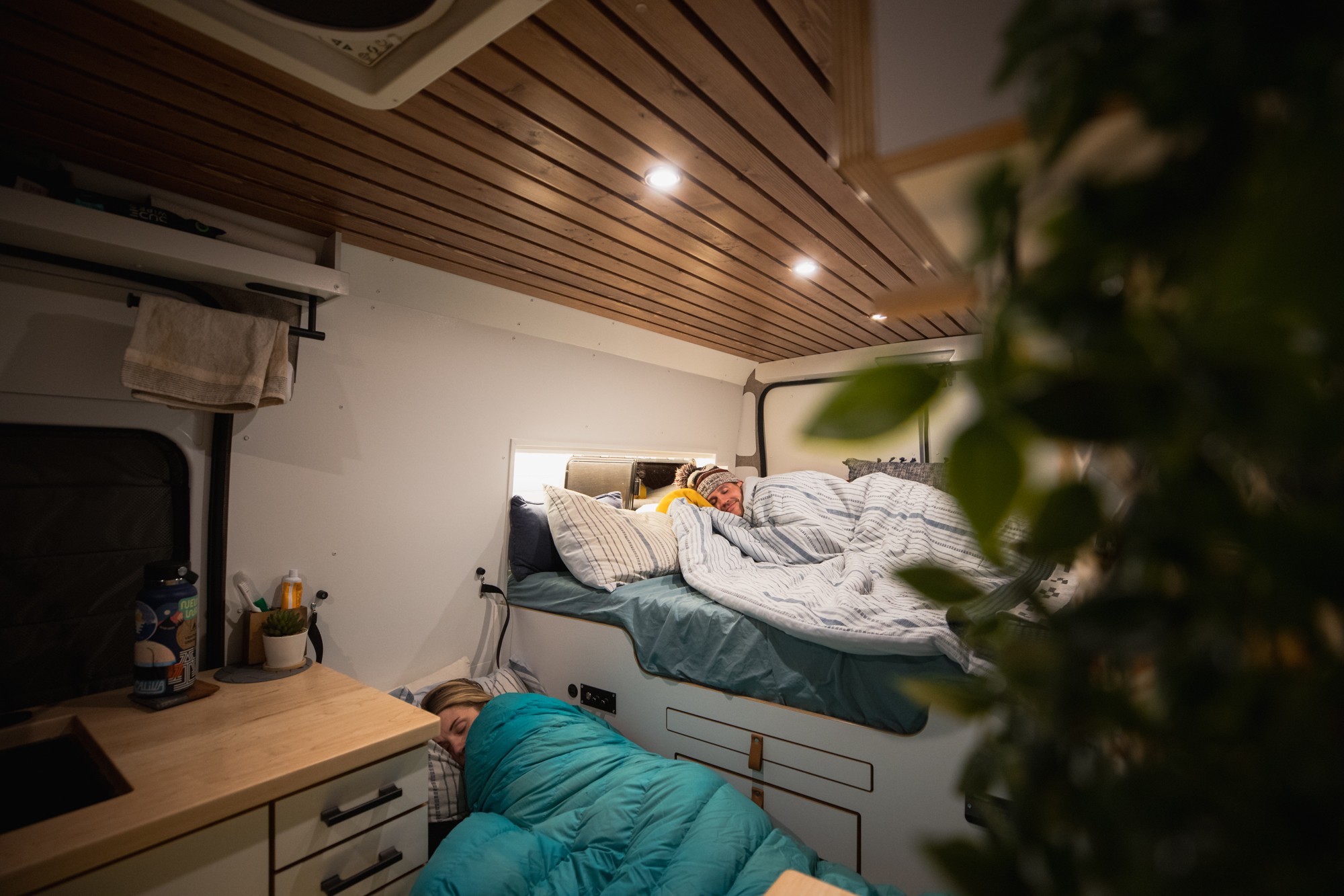 Inside a cozy RV rental: two people sleeping in a modern camper van with wood ceiling & warm lights. Ideal for adventures.. Dodge Ram Promaster 2021