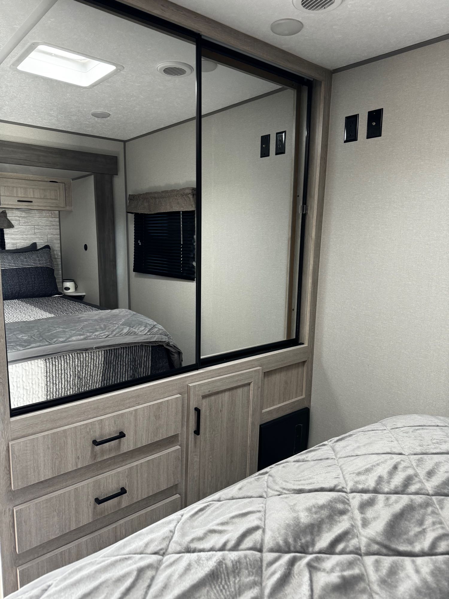 Modern RV rental bedroom features a mirrored closet, light wood cabinetry with drawers, and cozy gray bedding. Includes outlets and skylight.. Forest River Aurora 2024