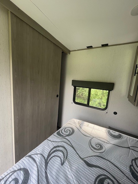 Compact RV rental bedroom interior features a patterned bed, small window with nature views, and a wooden sliding closet door.. Dutchmen Coleman Lantern 2022