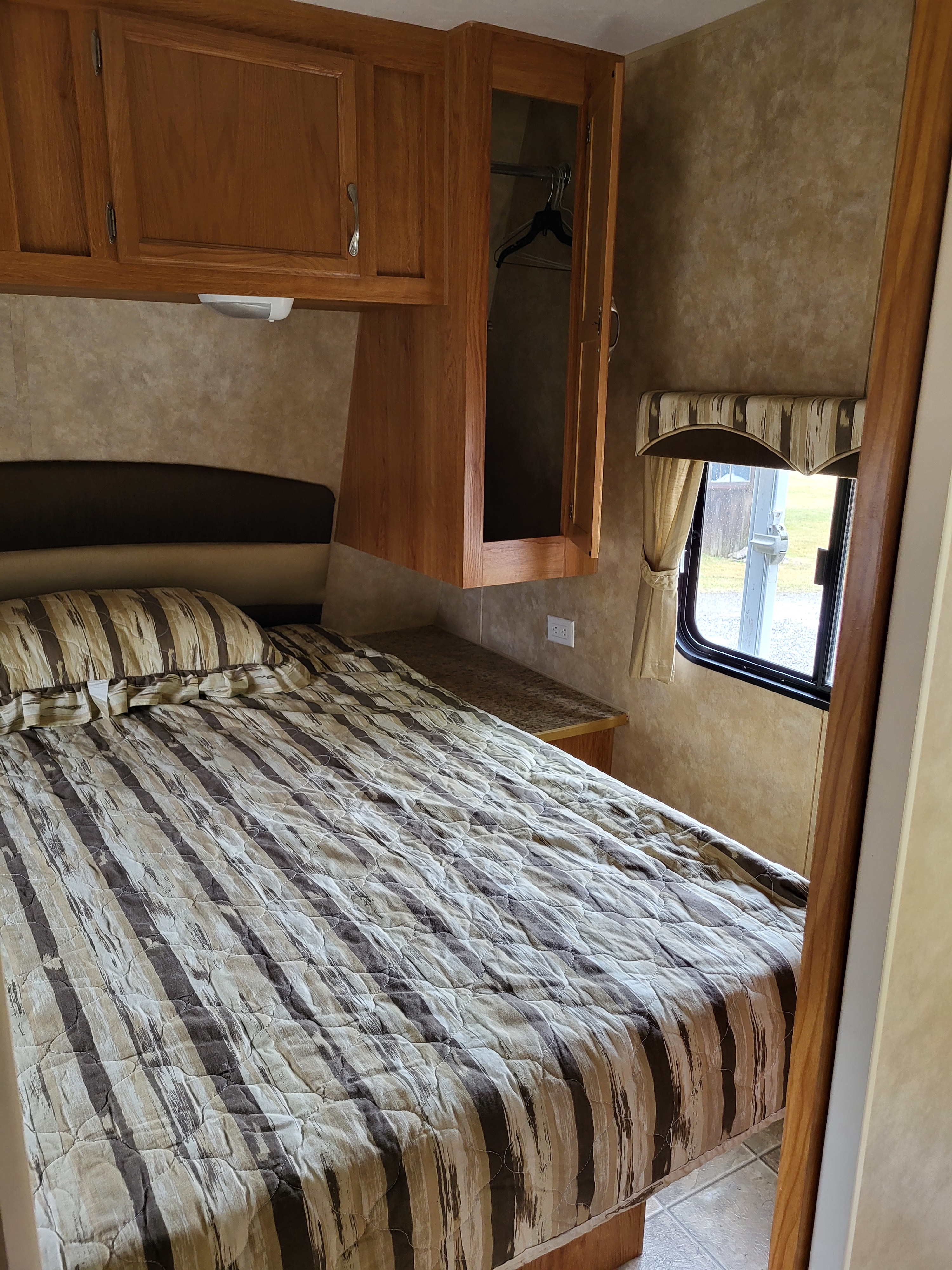 Cozy RV rental bedroom: striped bed, open overhead cabinet with hangers, and a side window.. Jayco Jay Flight 2008