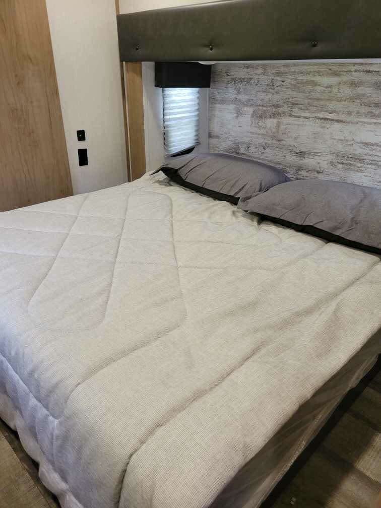 This cozy RV rental bedroom features a comfortable bed with gray pillows, a stylish textured wall, and a window blind.. Forest River Cherokee Arctic Wolf 2023