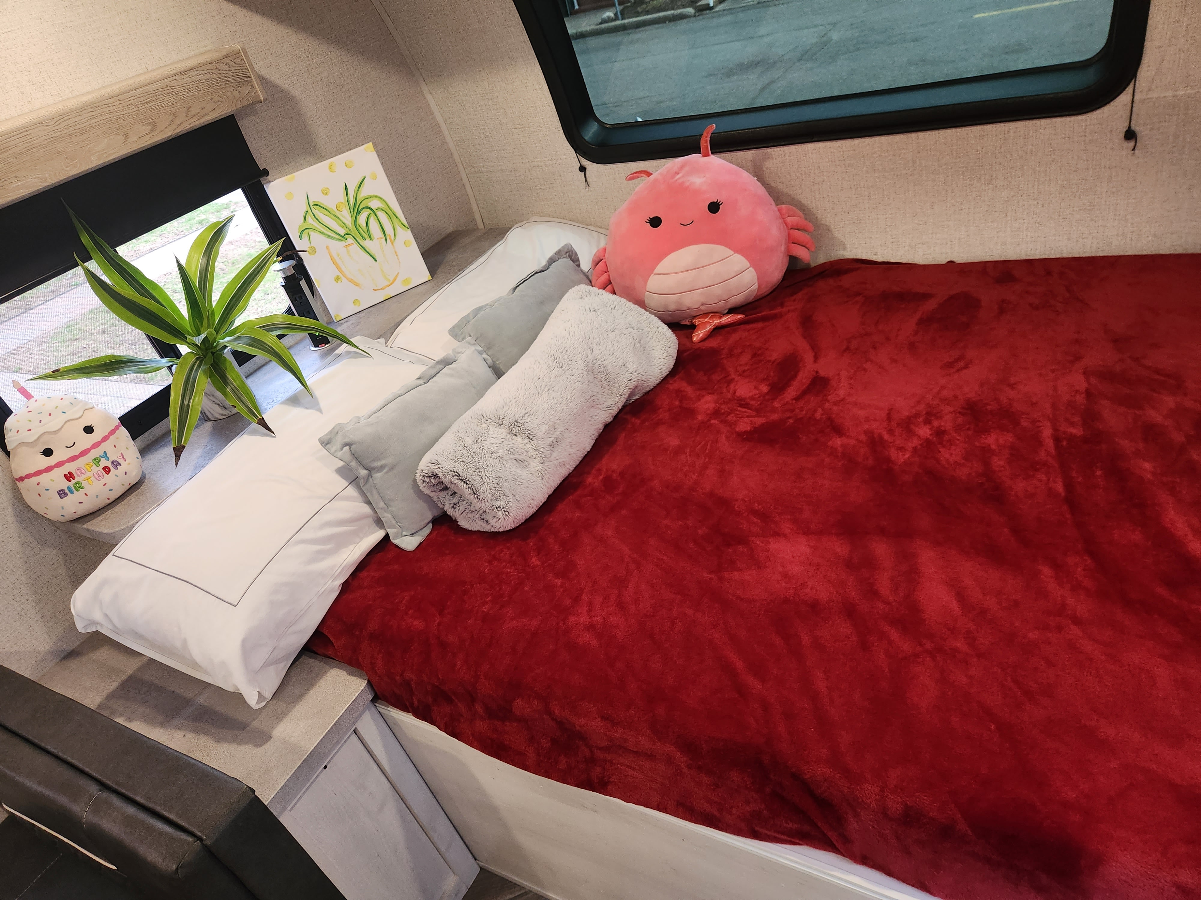 Cozy RV rental bedroom with a vibrant red blanket, plush pillows, and charming decor including a plant and window view.. Dutchmen Coleman Rubicon 2021