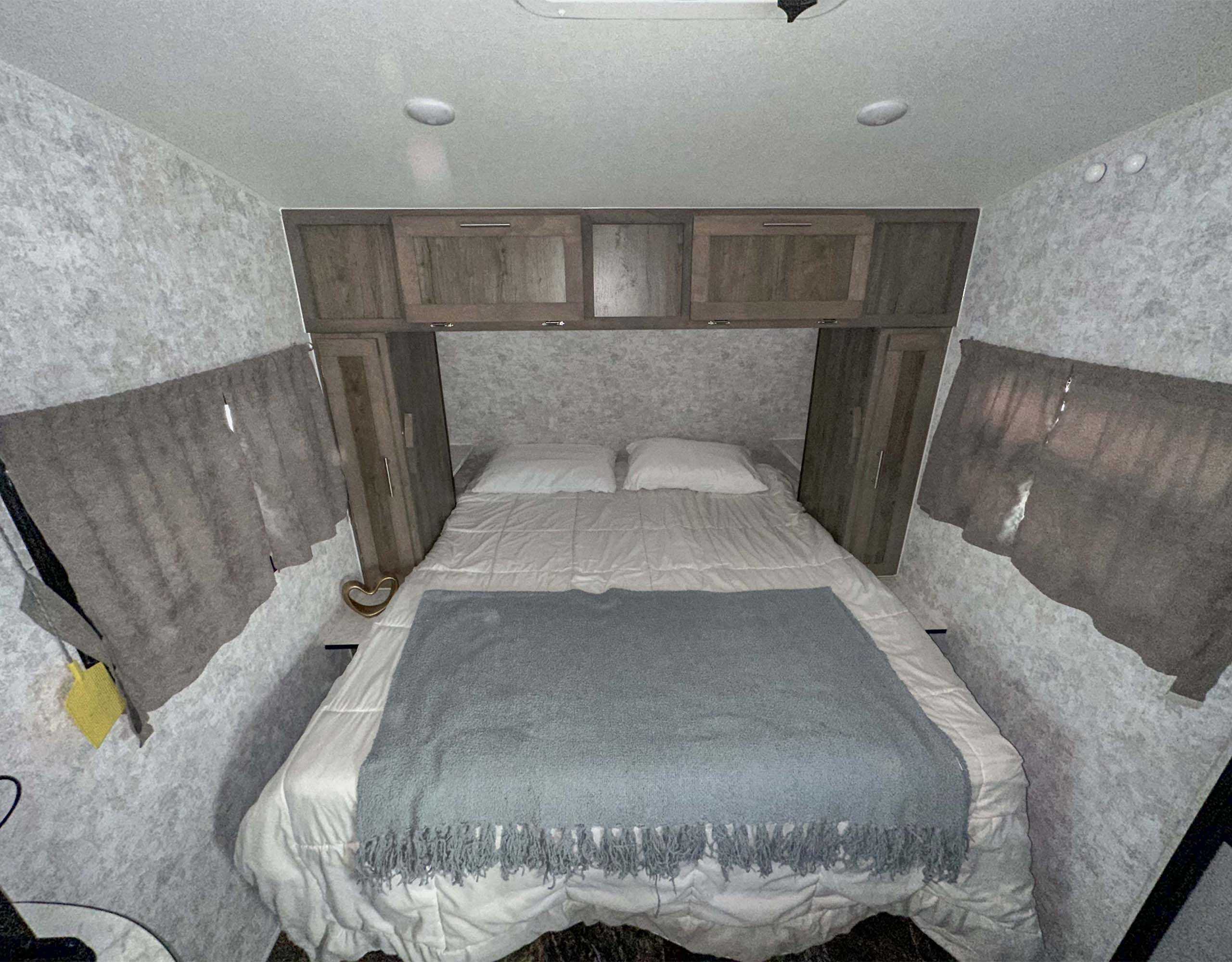 Functional RV rental bedroom with bed, white bedding, grey throw, and overhead cabinets. Features light textured walls and side windows.. Prolite Classic 2023