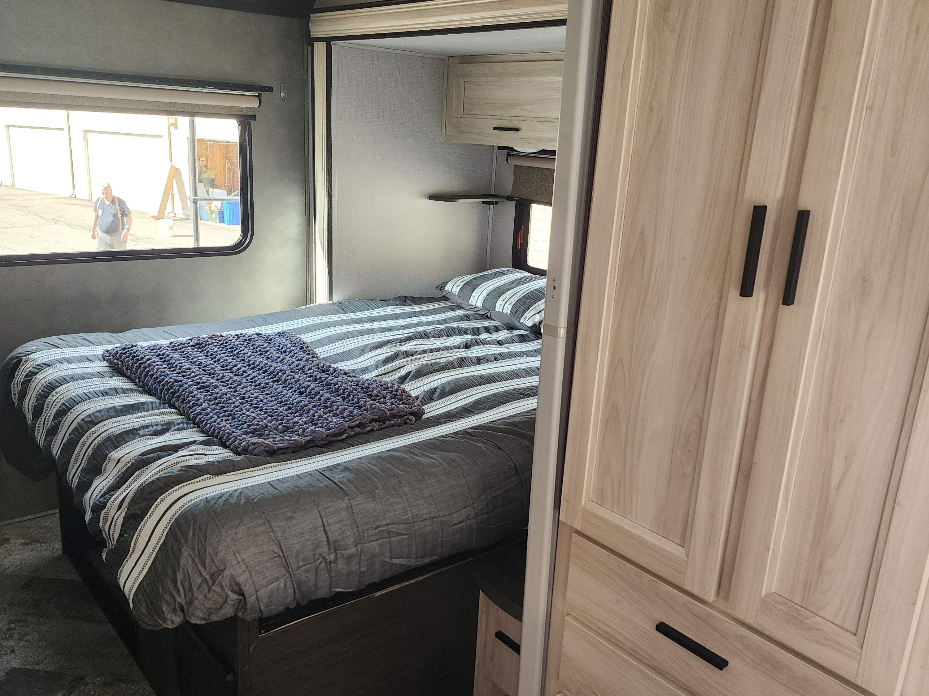 Cozy RV rental bedroom with a striped bed, knitted blanket, light wood storage, and a person visible outside the window.. Forest River Forester 2020