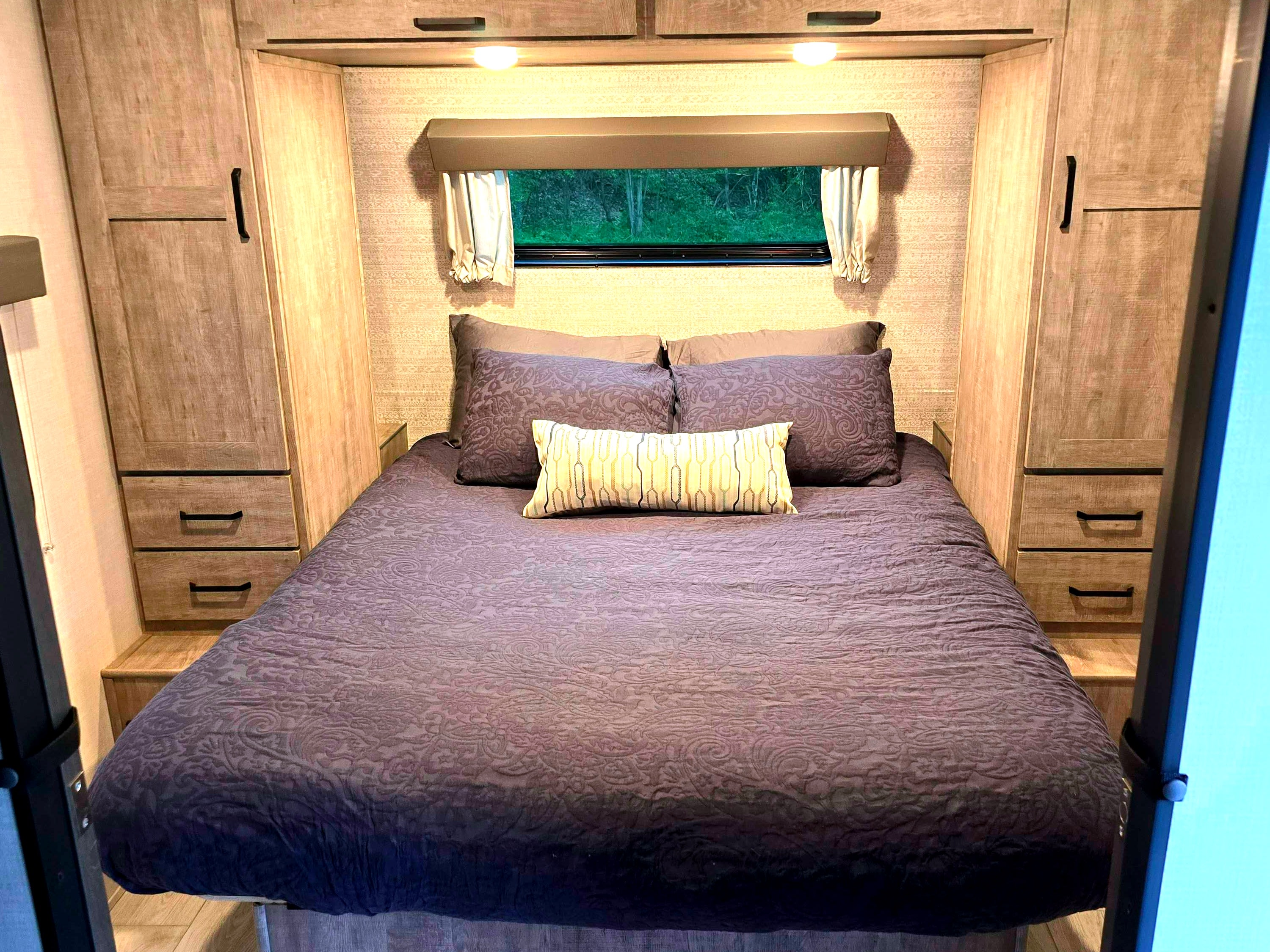 Cozy RV rental bedroom featuring a queen bed with purple bedding, built-in wooden storage, and a window overlooking nature.. Forest River Surveyor Legend 2023